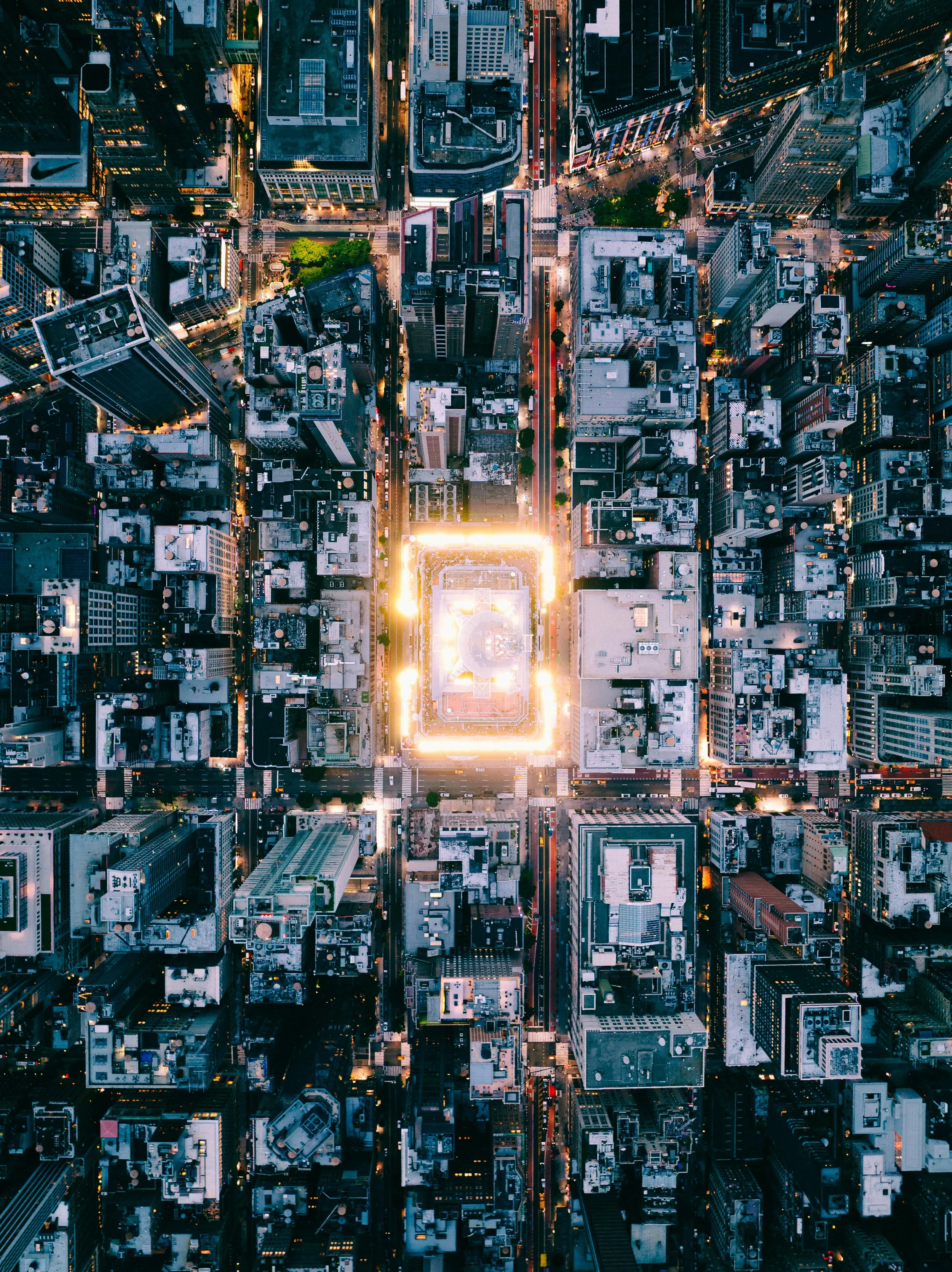 Aerial view of a city at dusk showing illuminated streets, skyscrapers, and a central rectangular building with bright lights.