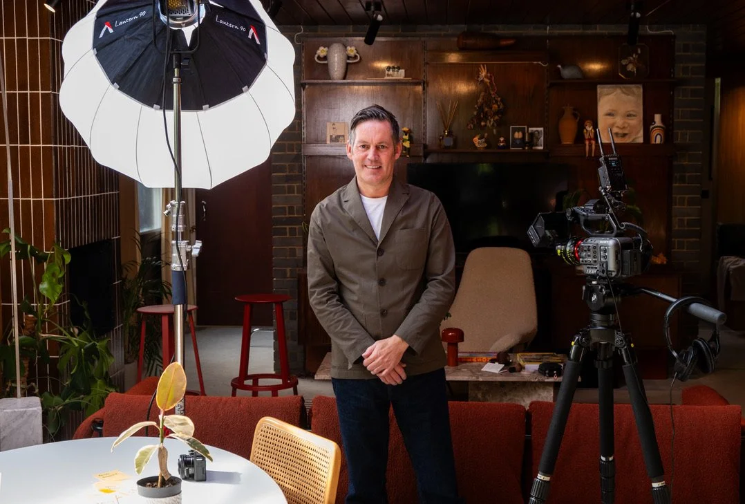 A man standing in a cozy, warmly lit living room or studio with professional photography equipment including a large softbox light and a camera on a tripod. The room features a wooden wall with decorative items, a television, and a framed photo of a smiling child.