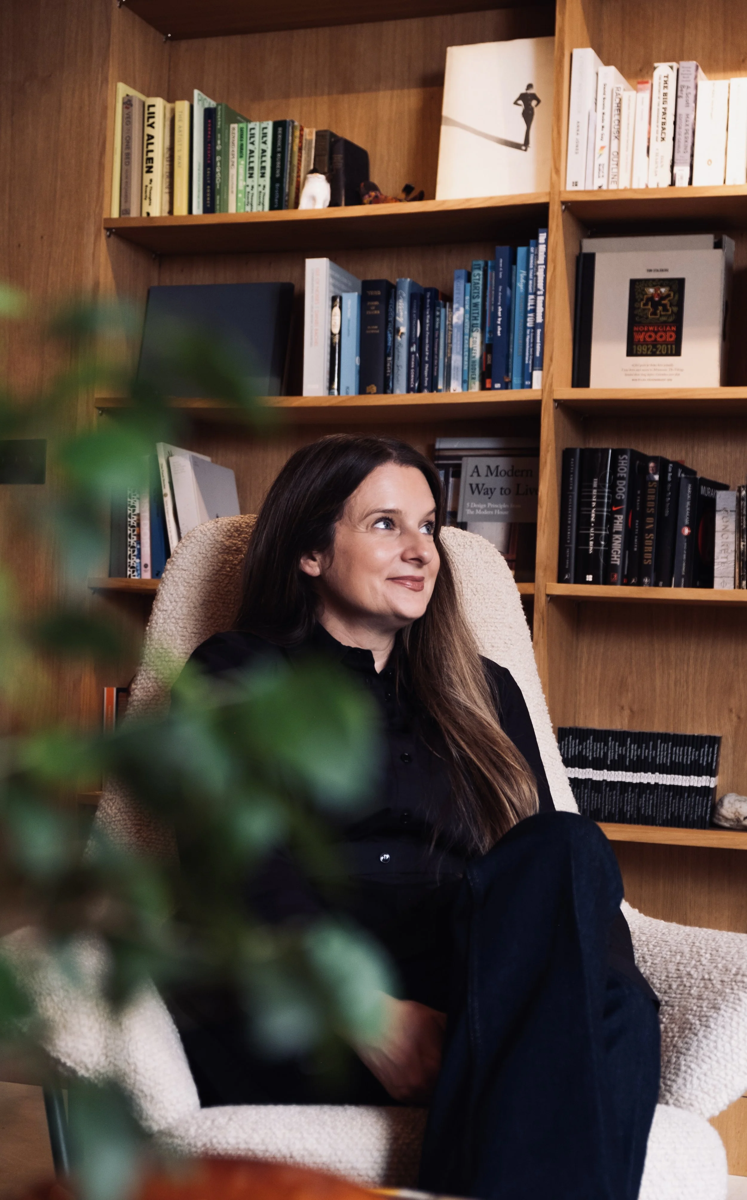 A woman sitting in a beige armchair in front of a wooden bookshelf filled with books, looking to the side with a slight smile.