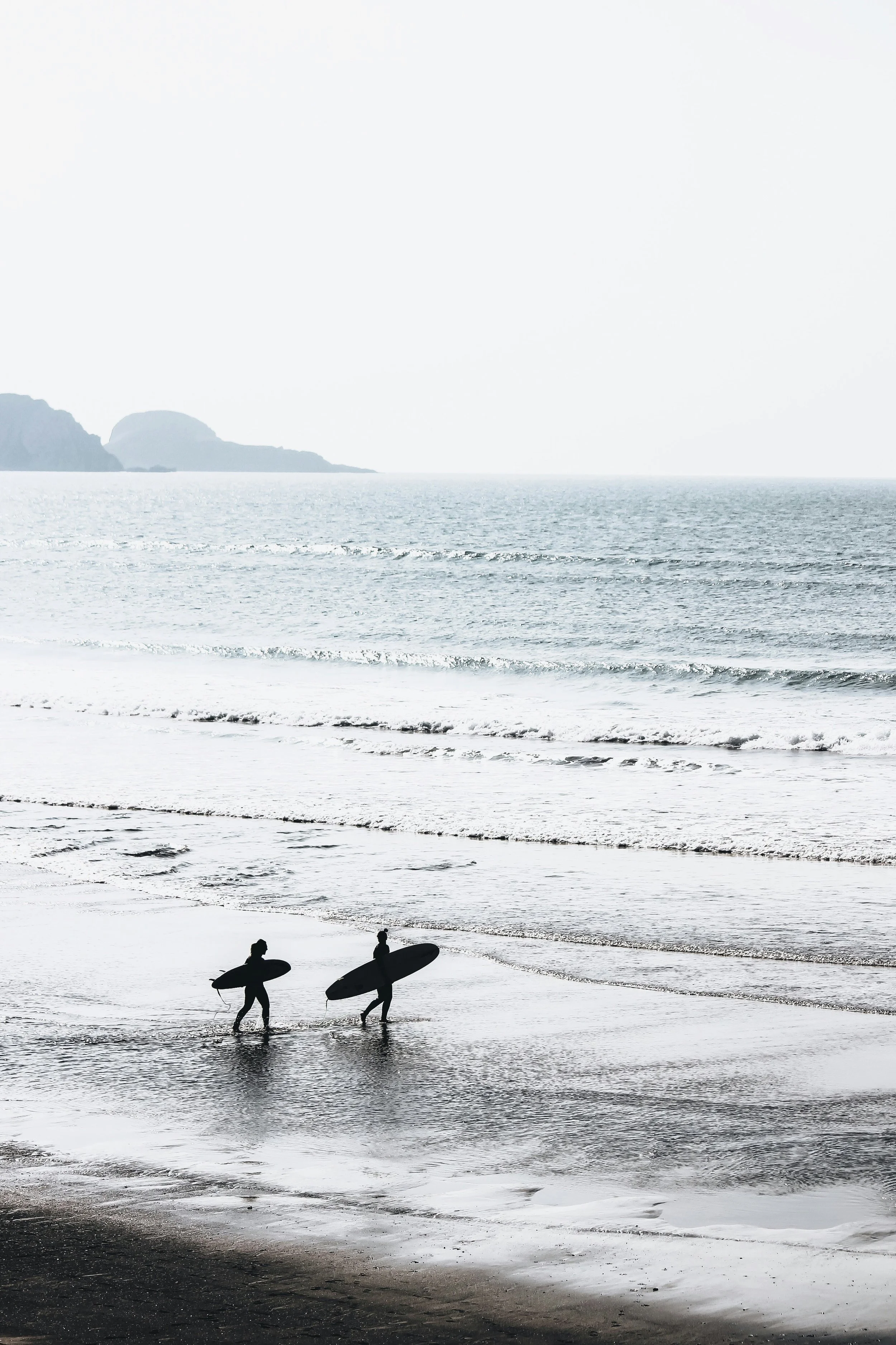 Two surfers walking along the beach with surfboards, approaching the water as waves roll in, in a black and white photo.