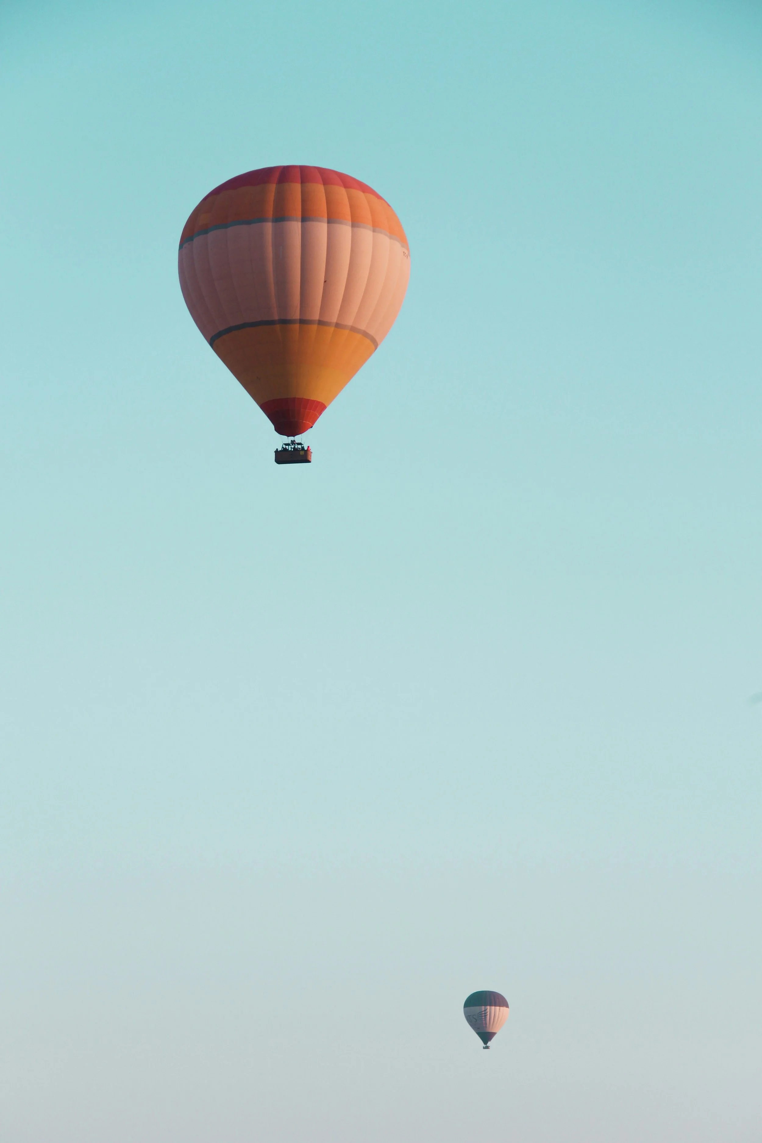 Two hot air balloons floating in a clear sky, with one large orange and peach balloon in the foreground and a smaller balloon in the background.