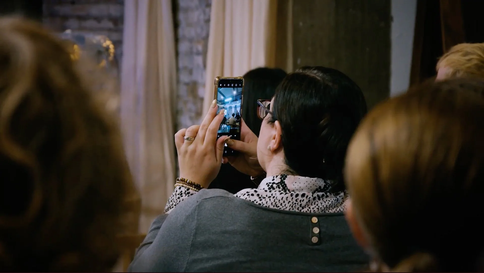 A woman with black hair, glasses, and bracelets is taking a photo with her smartphone at a gathering or event, surrounded by other people.