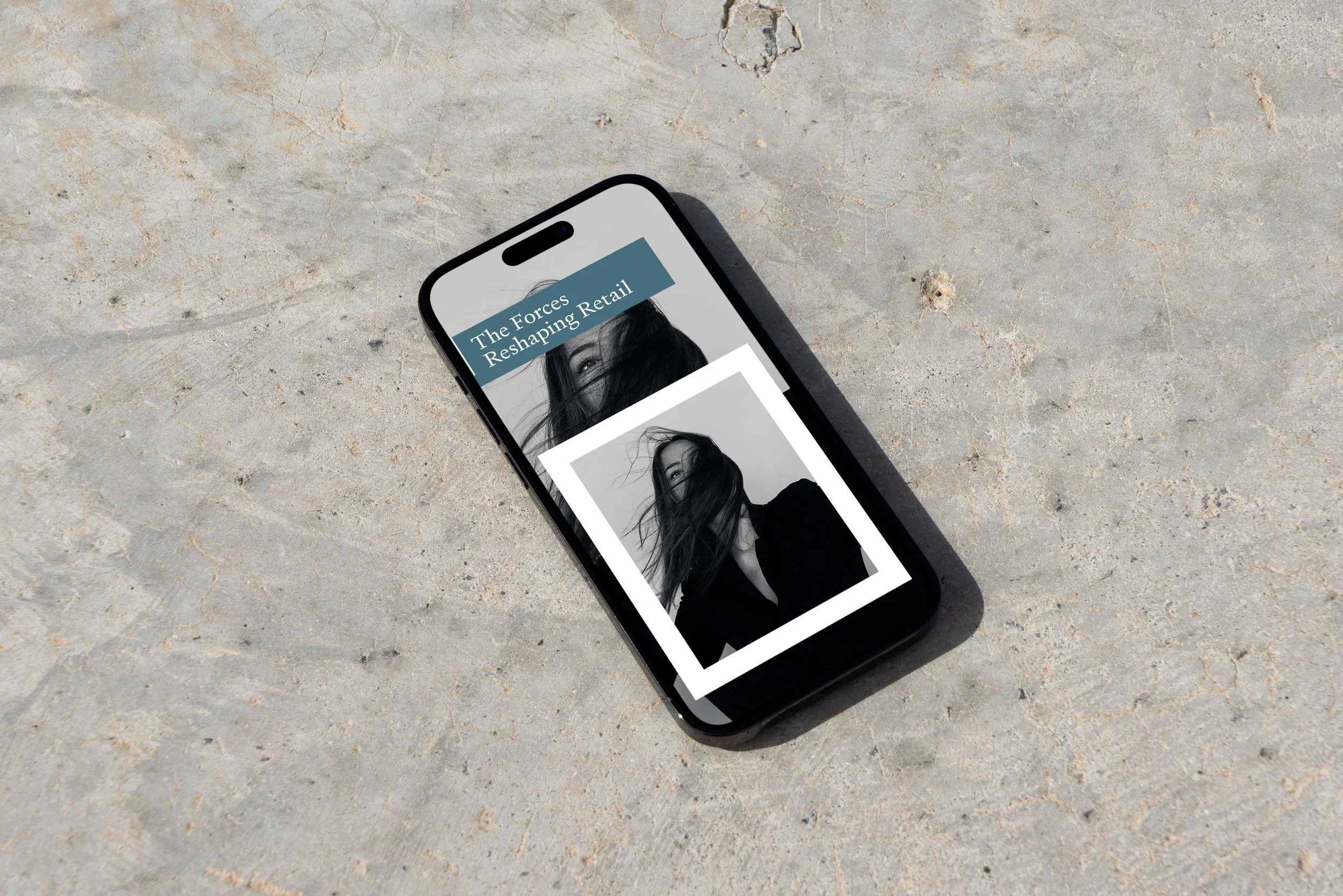 A smartphone lying on a sandy surface displaying an article titled "The Forces Reshaping Retail" with a black-and-white portrait of a woman with hair covering part of her face.
