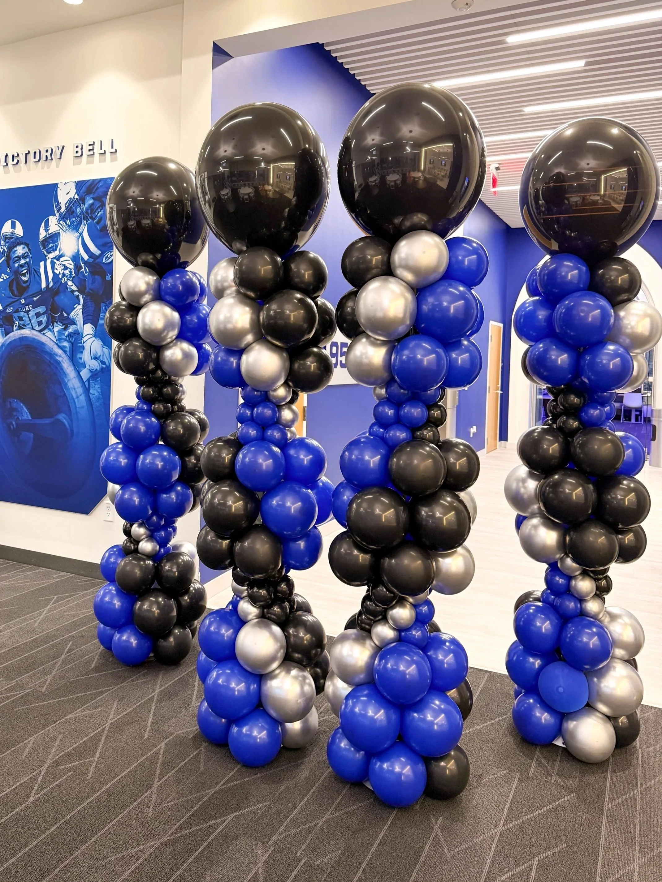 Four columns of balloons in black, blue, and silver, topped with large black balloons reflecting the room interior, at a sports facility with a blue wall and sports-themed artwork in the background.