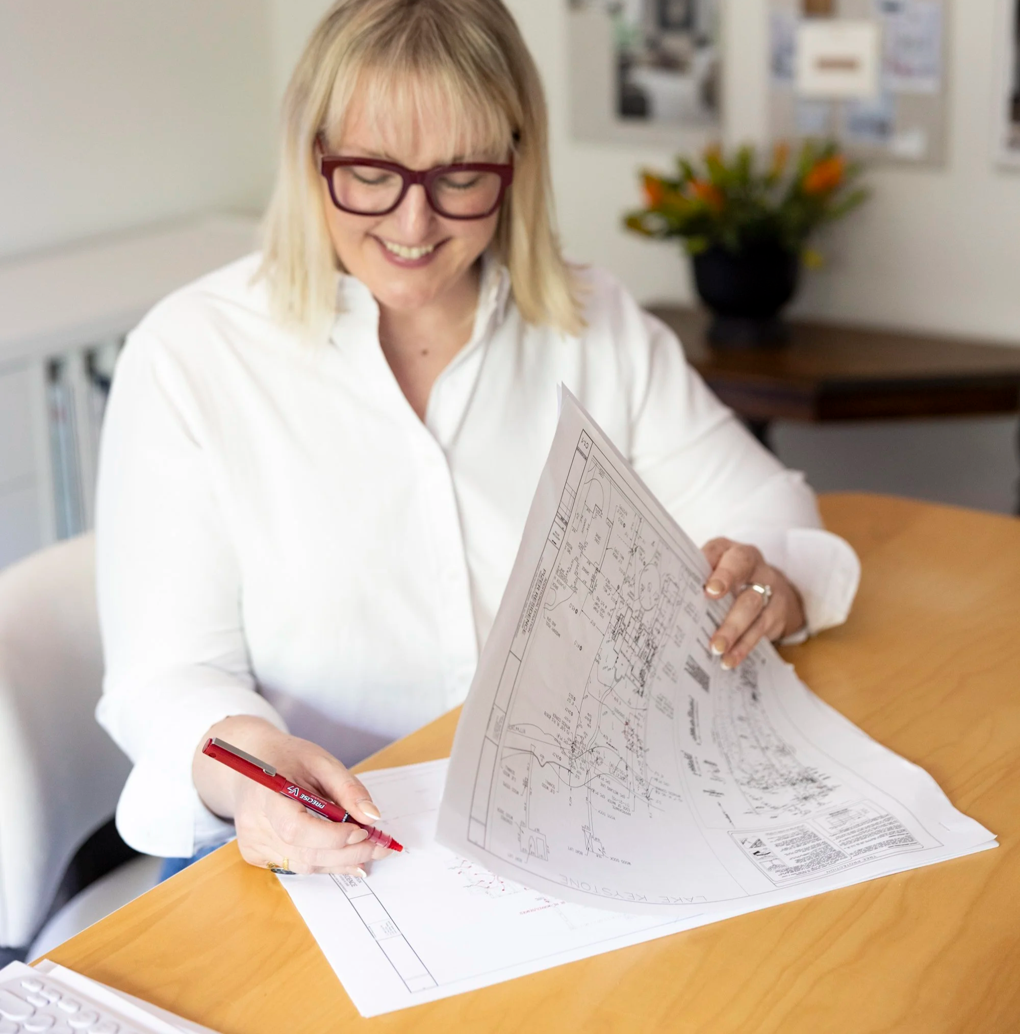 Woman with blonde hair and glasses smiling while working with architectural plans at a desk, with a plant in the background.