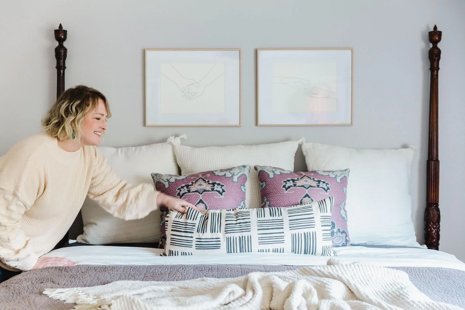 A woman with short blonde hair, wearing a cream sweater, smiling and adjusting decorative pillows on a bed with white and purple pillows. The background features two framed minimalist line drawings of hands and a bed with wooden posts.