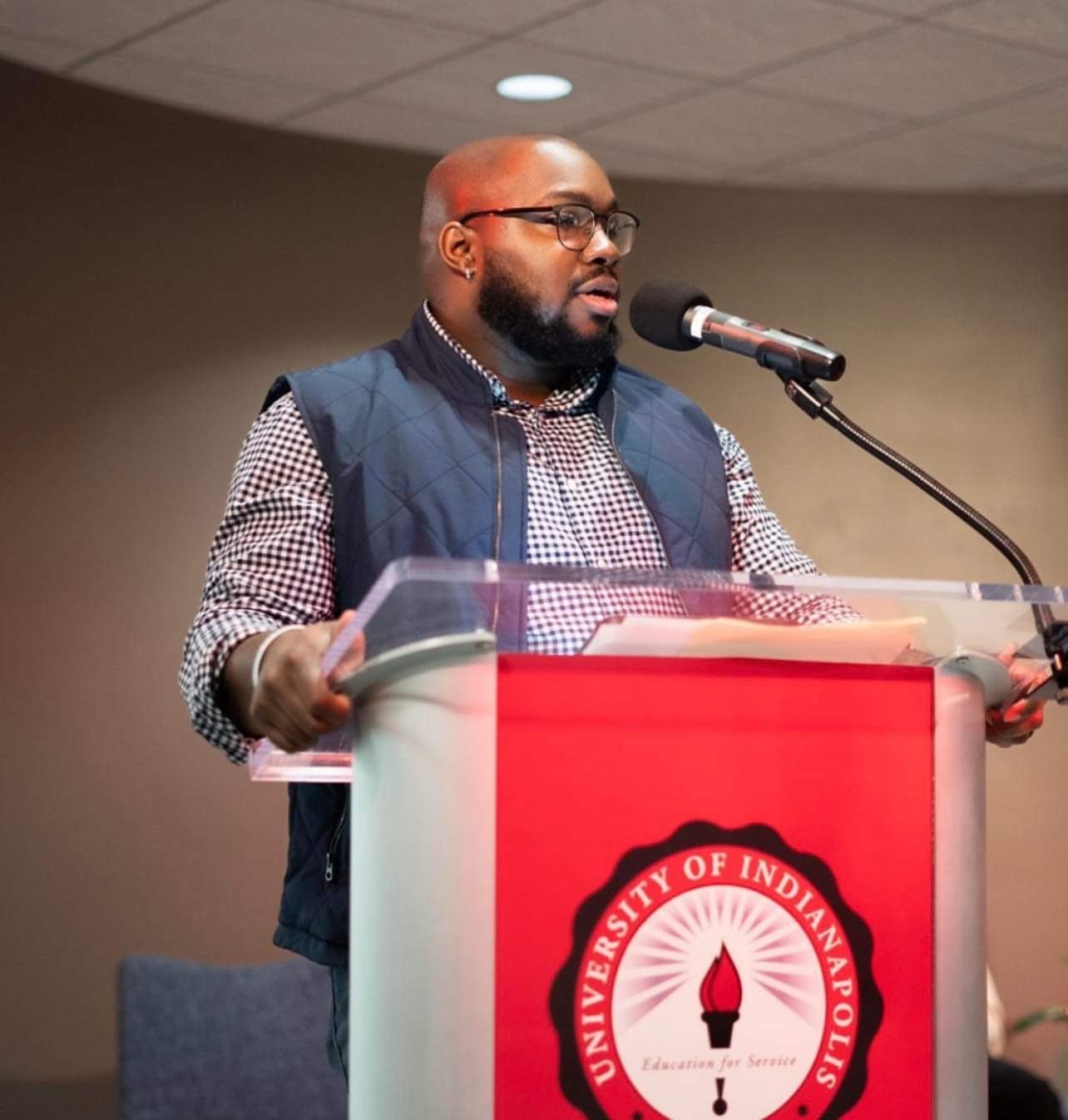 Thomas Kneeland standing at a podium, at the University of Indianapolis, wearing glasses, a checkered shirt, and a navy vest, with a microphone in front of him.