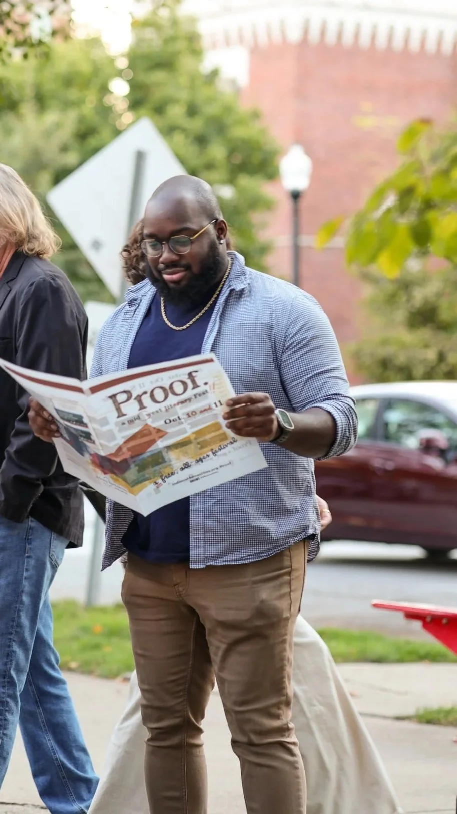 Thomas Kneeland, wearing glasses and a blue checkered shirt with brown pants, reading a newspaper featuring Proof: A Midwest Lit Fest outdoors with trees, a lamp post, and with Indiana Humanities in the background.