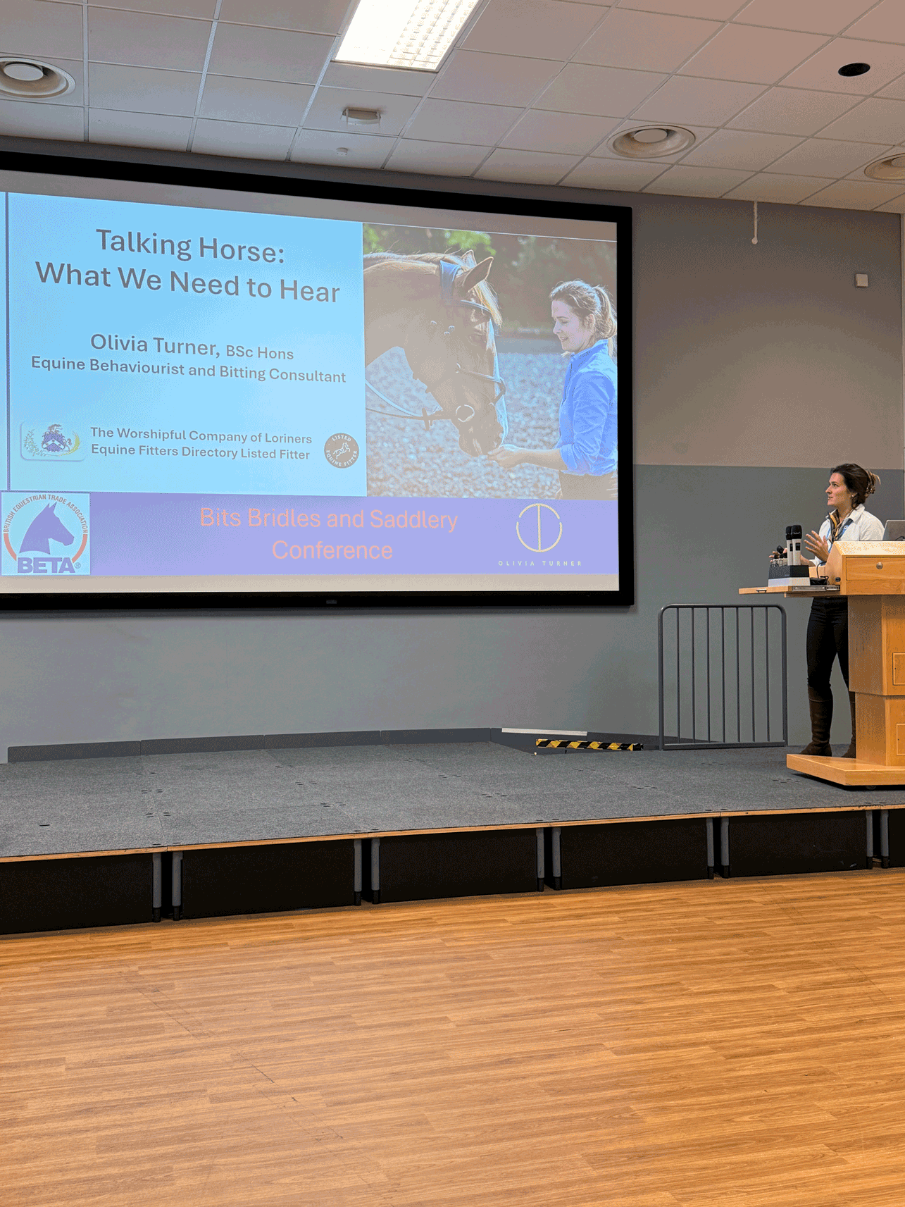 A woman giving a presentation next to a large screen displaying a slide about talking horses, with a photograph of a woman and a horse, and text including "Talking Horse: What We Need to Hear," and logos of equestrian organizations.