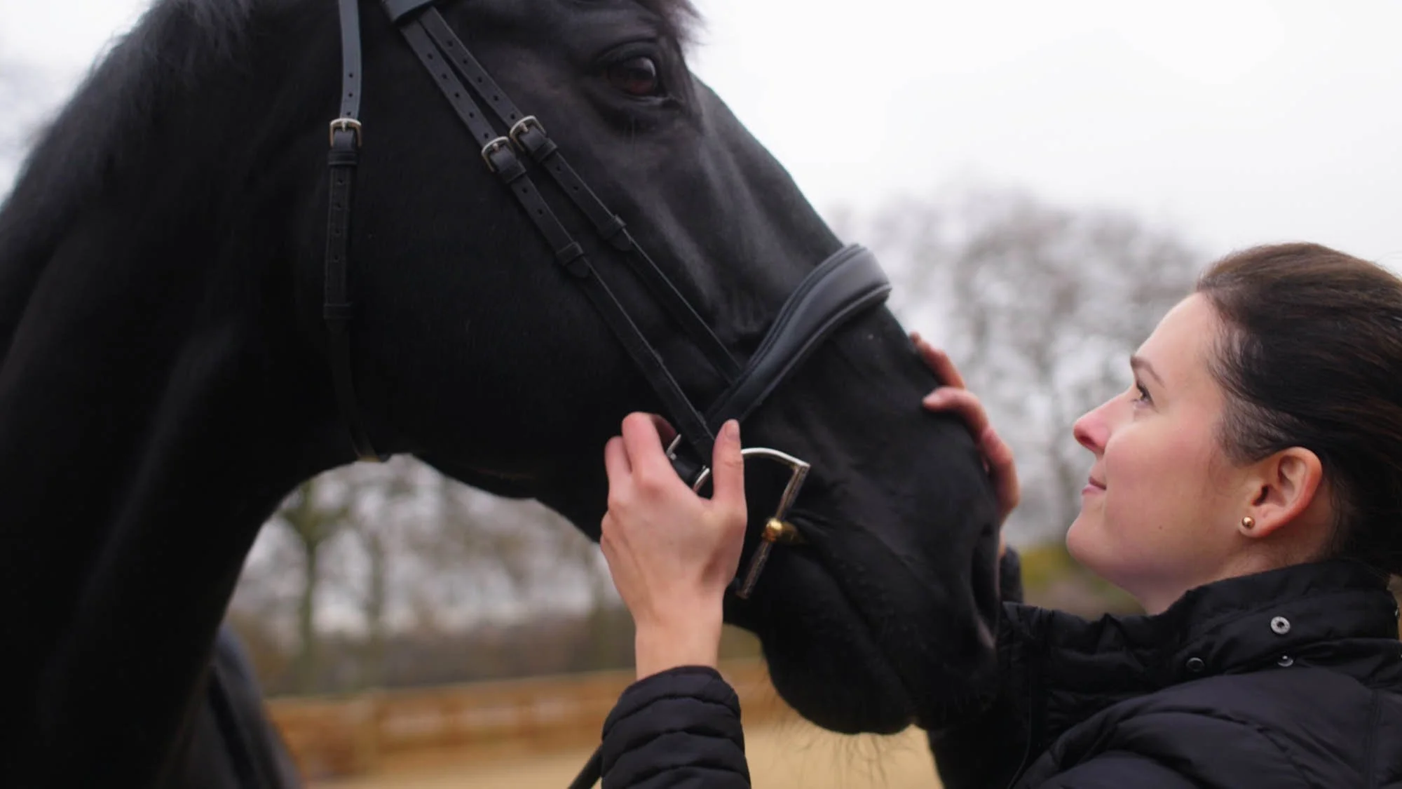 A woman lovingly gazes at a black horse, gently touching its face outdoors on a cloudy day.