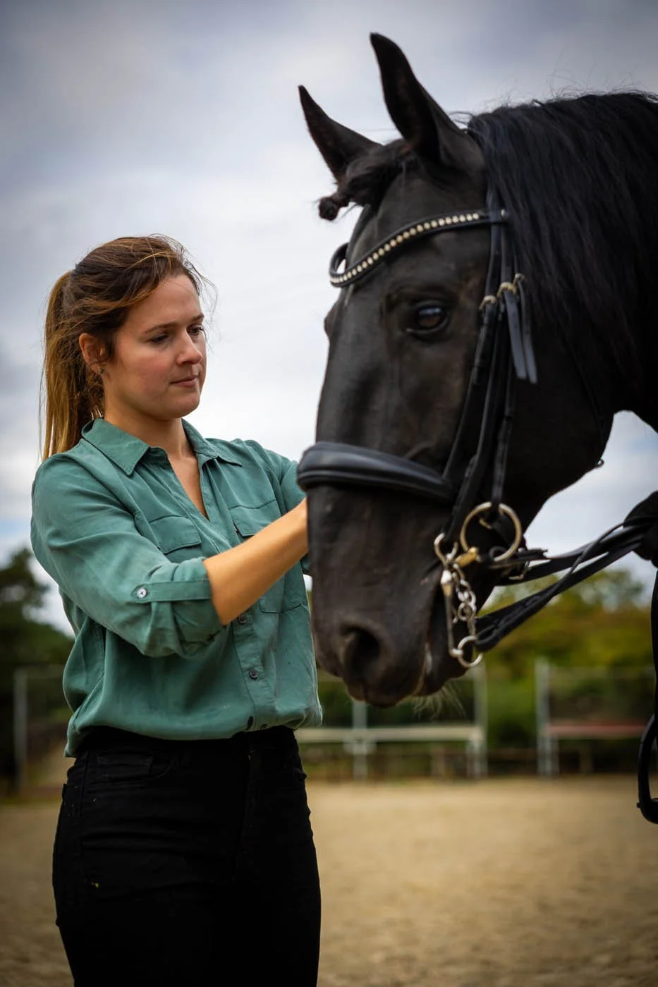 A woman is adjusting the bridle of a black horse with a dark sky in the background.