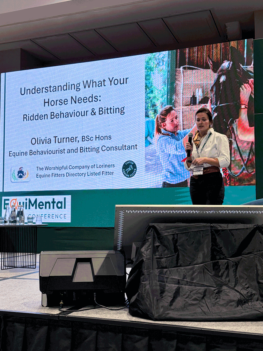 A woman in a white shirt and black pants standing on a stage, speaking into a microphone at a conference. Behind her, a large screen displays a presentation slide with the title 'Understanding What Your Horse Needs: Ridden Behaviour & Bitting' by Olivia Turner, an equine behaviourist and bitting consultant. The slide features images of a woman and a horse. There are water bottles on a table on the stage, and the background shows a large conference room with a black curtain.
