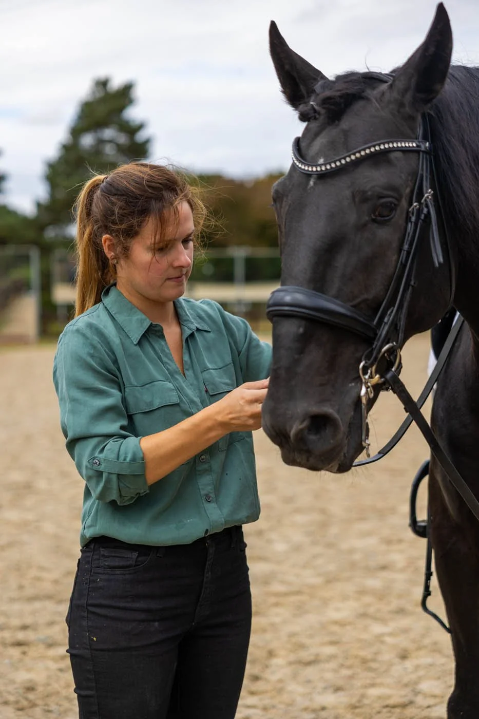 A woman in a teal shirt adjusting the bridle of a black horse in an outdoor riding arena.