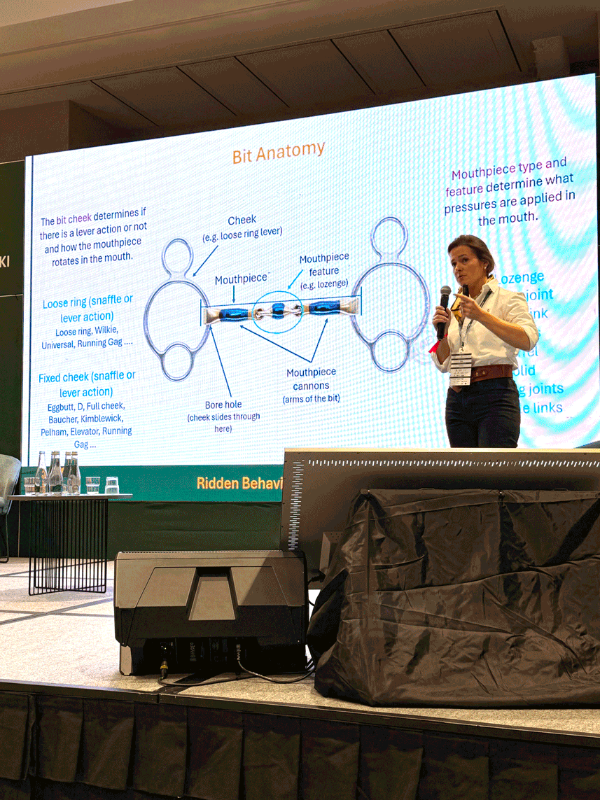 A woman giving a presentation on bit anatomy, with a slide displaying labeled parts of a bit, including cheek, mouthpiece, cannons, bore hole, and pressure features, at a conference or seminar.