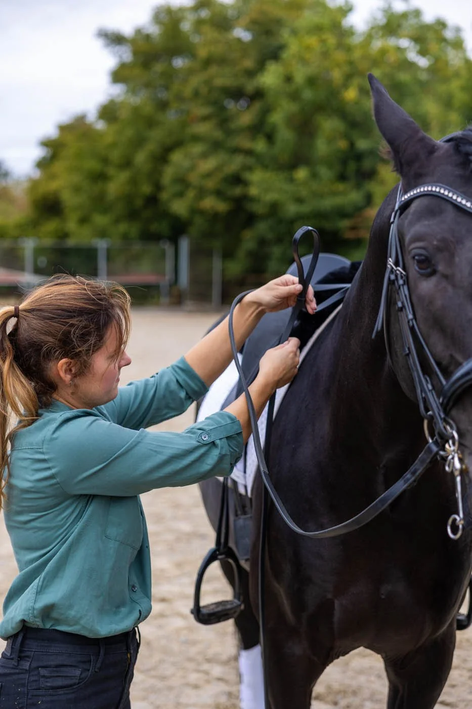 A woman is adjusting the saddle or bridle on a black horse in an outdoor riding arena.