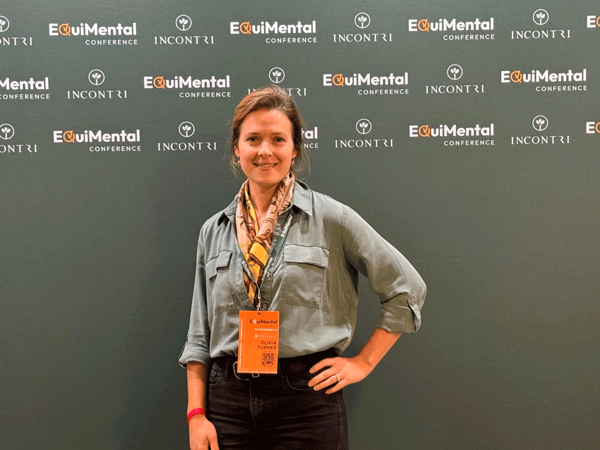 A woman standing in front of a black backdrop with logos for the EquiMental Conference and Incontri, smiling, wearing a gray shirt, black pants, a colorful scarf, and an orange conference badge.