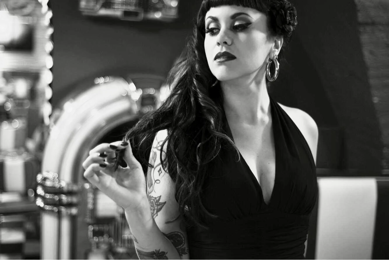 A woman with tattoos in a black dress looks to the side, holding her hair in a retro diner setting, black and white photo.