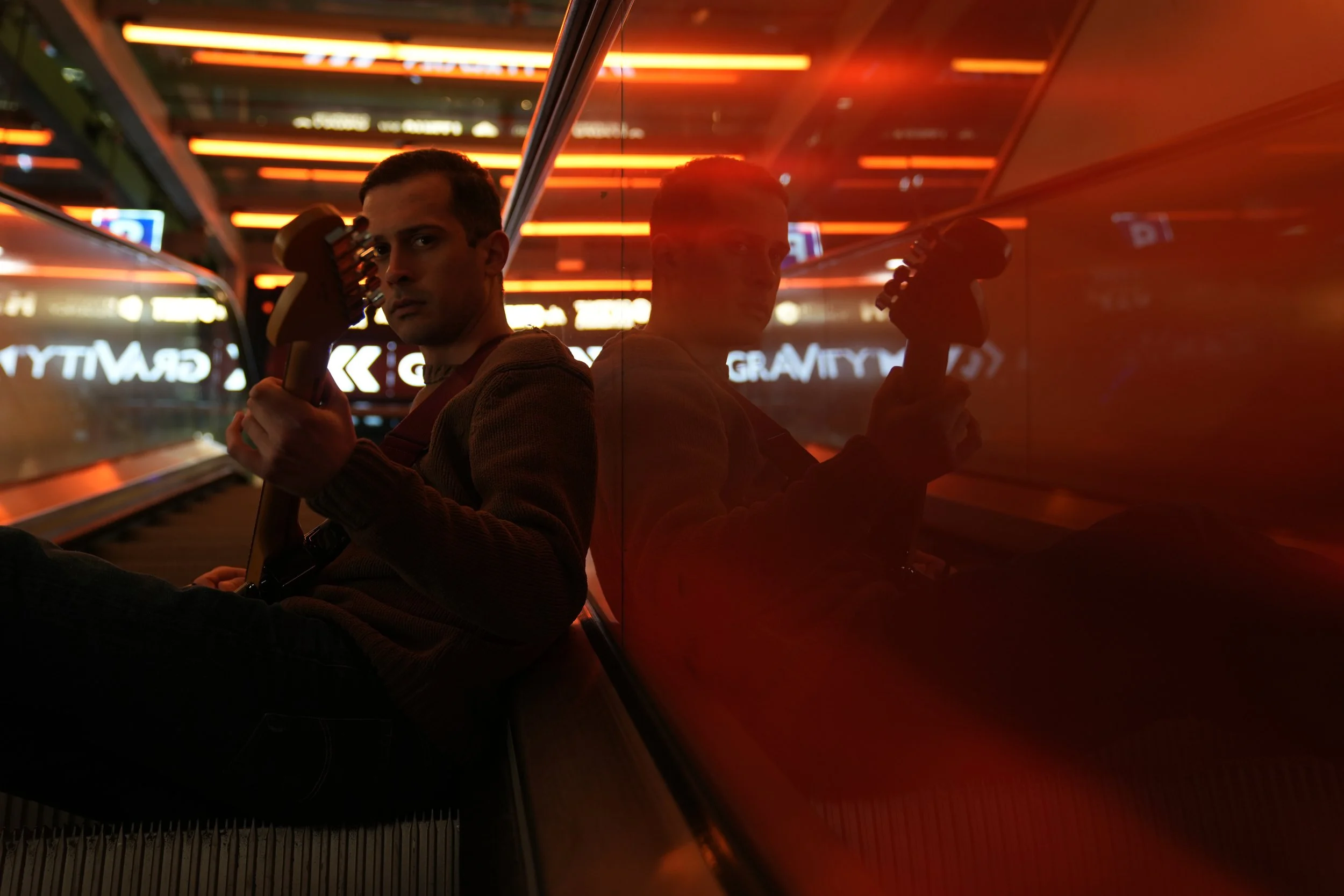 A man holding a guitar sits on an escalator, his reflection visible on the glass beside him under orange lights.