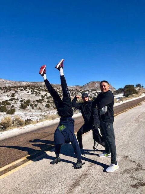 Three men in black jackets and pants, two of them standing and one doing a handstand on the road. The handstand man is wearing red sneakers and has his legs held straight up. The other two men are smiling, with one holding the handstand man's ankle. 