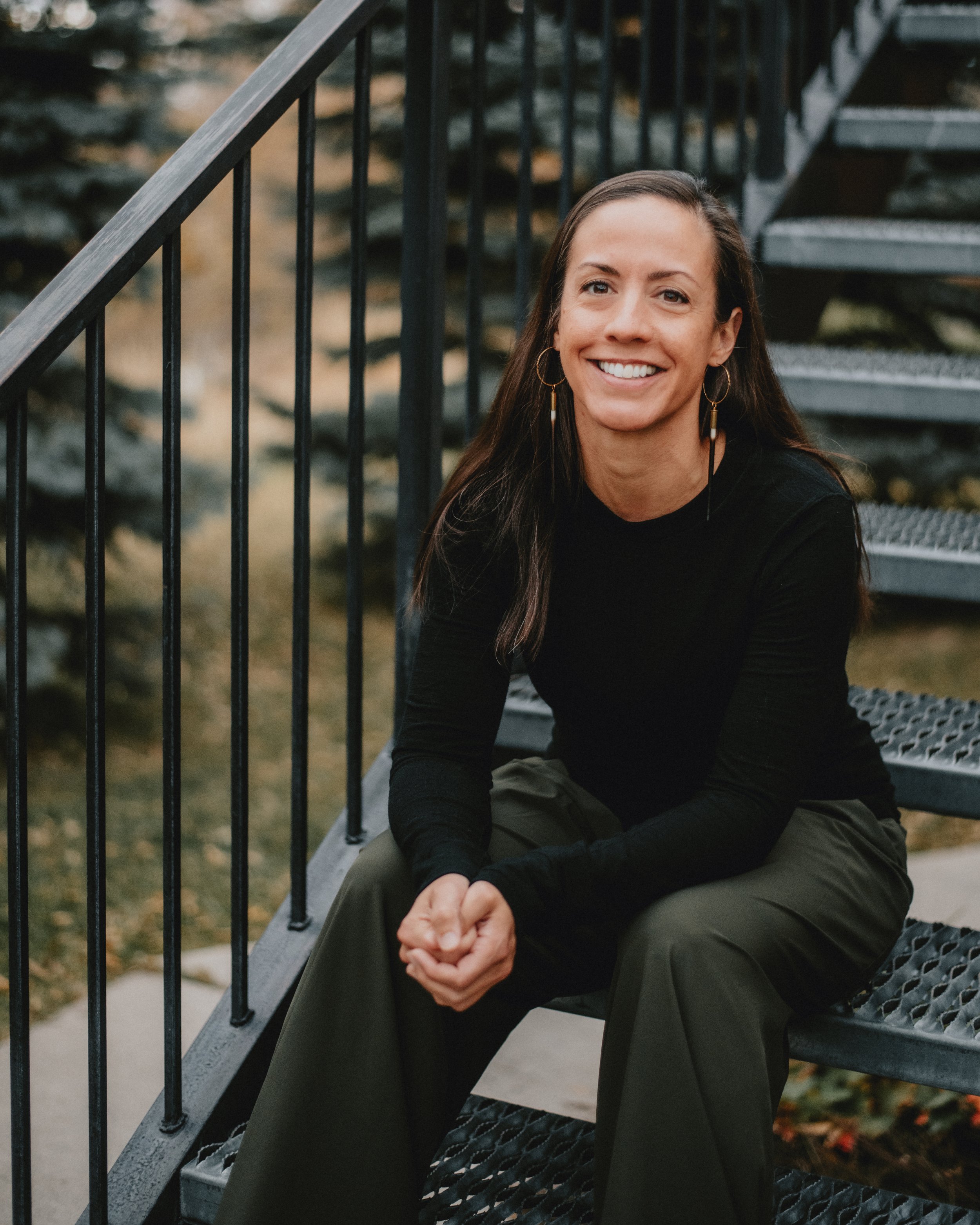 Image depicts therapist Erin Good sitting on a metal staircase. Erin is smiling and looking at the camera. She is dressed in a black long sleeve shirt and olive green pants.
