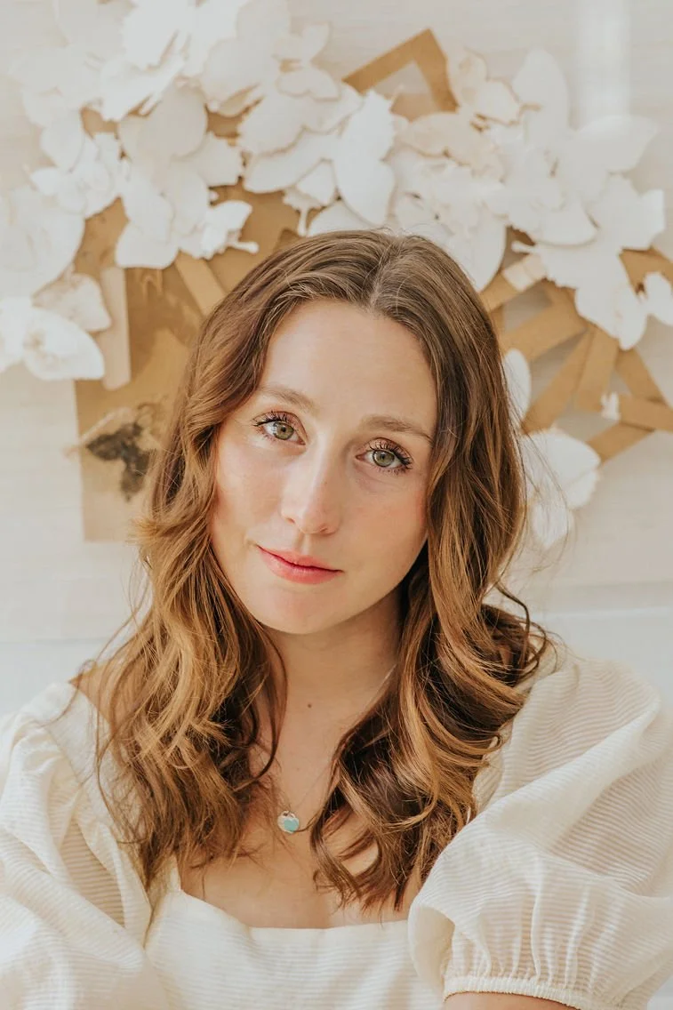 Portrait of a young woman with wavy brown hair wearing a cream-colored top, standing in front of a white and wooden decorative art piece with paper butterflies.