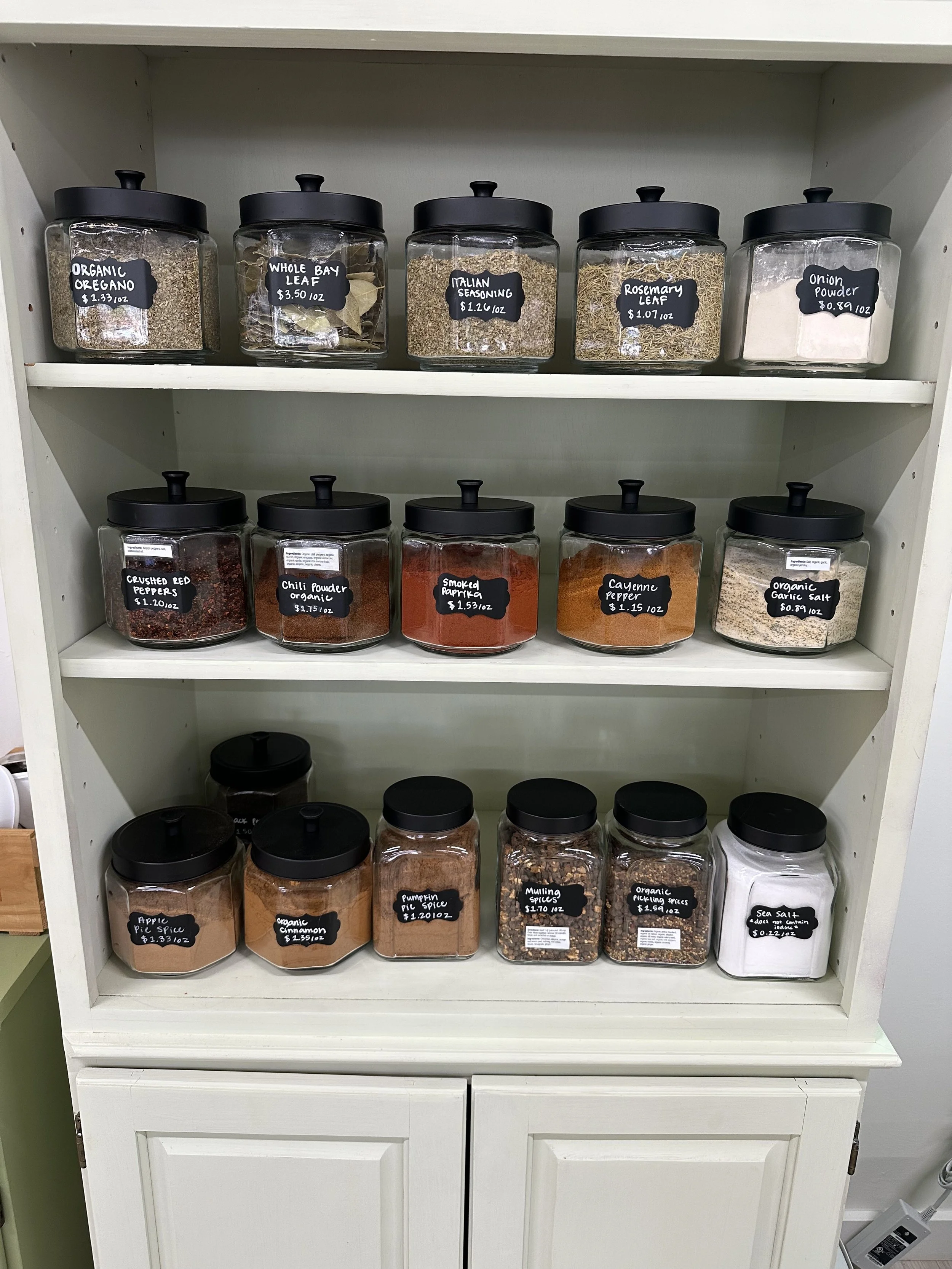 White shelves with glass jars filled with spices and herbs, with black labels and lids. The spices include oregano, bay leaf, Italian seasoning, rosemary leaf, onion powder, crushed red peppers, chili powder, paprika, garlic salt, apple pie spice, cinnamon, pumpkin pie spice, mullina spices, and sea salt.