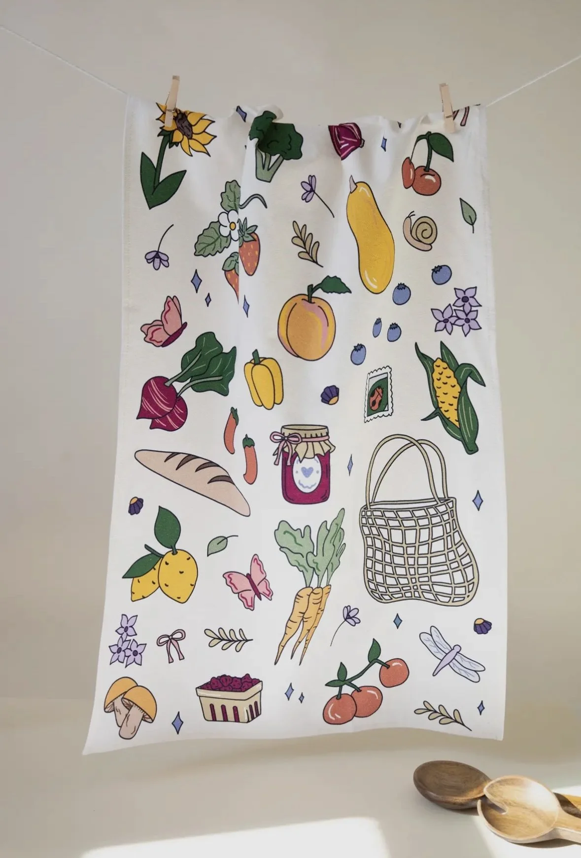 A white tea towel with colorful illustrations of fruits, vegetables, flowers, a jar of jam, a basket, a mushroom, and butterflies hanging on a line with clothespins, with some wooden spoons placed at the bottom right.
