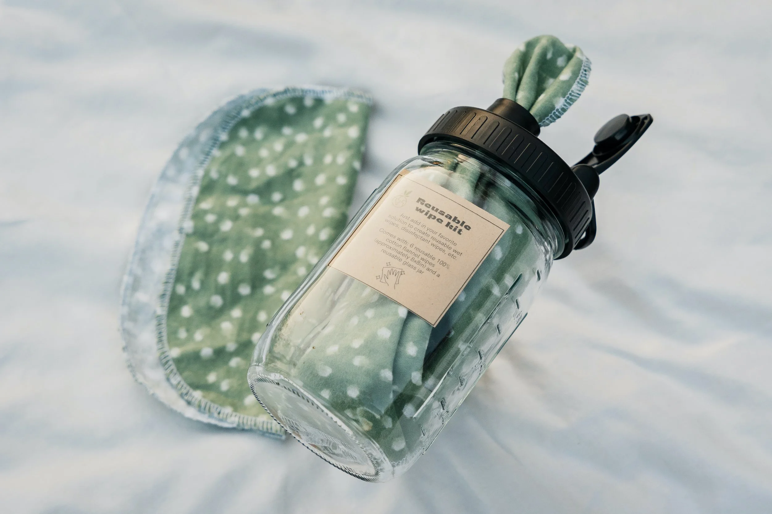 A glass water bottle with a black lid and a reusabale wipe belt, lying on a soft white surface with a green polka dot cloth nearby.