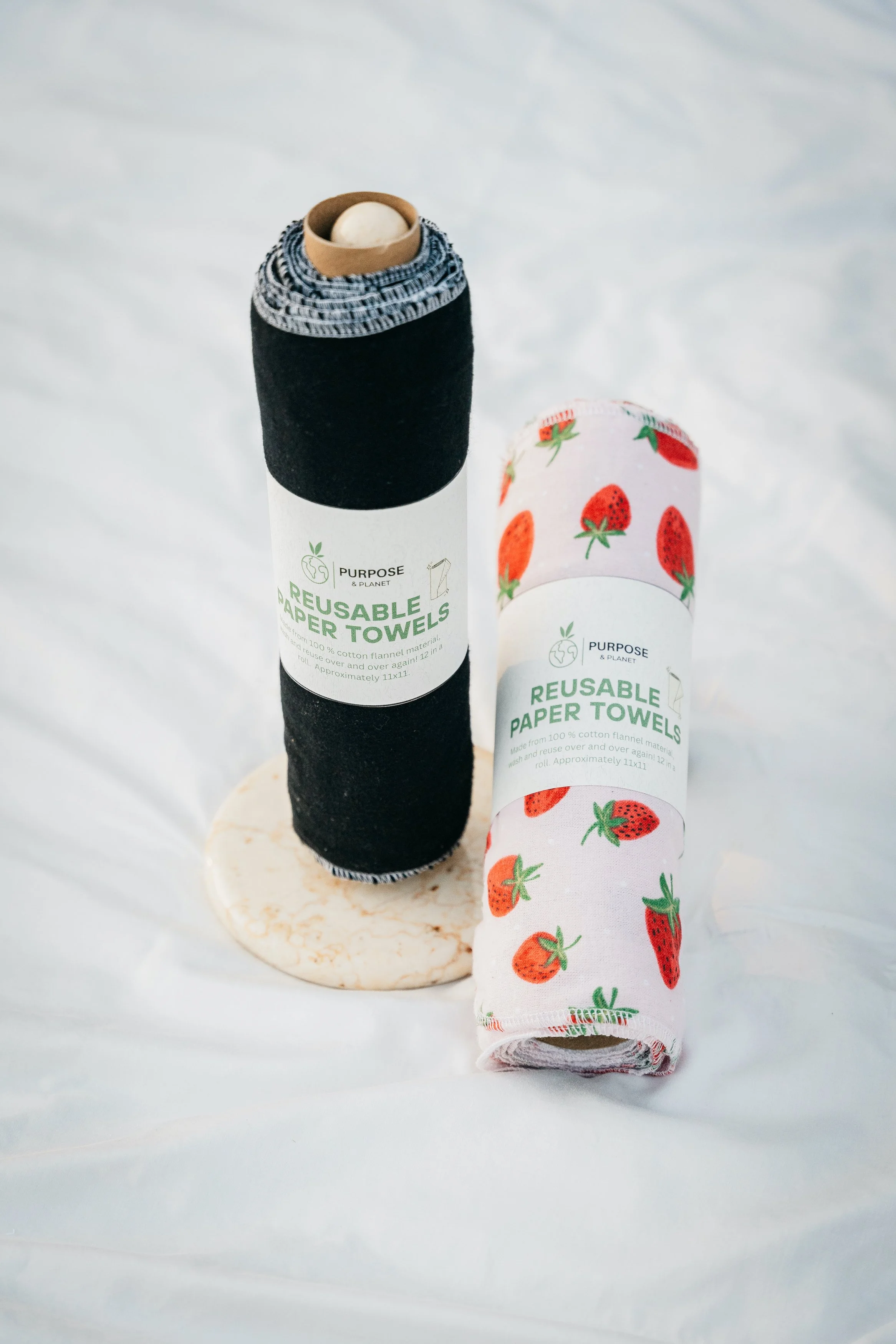 Two rolls of reusable paper towels, one with a black and white striped pattern and the other with a strawberry pattern, standing vertically on a small round stand against a white background.