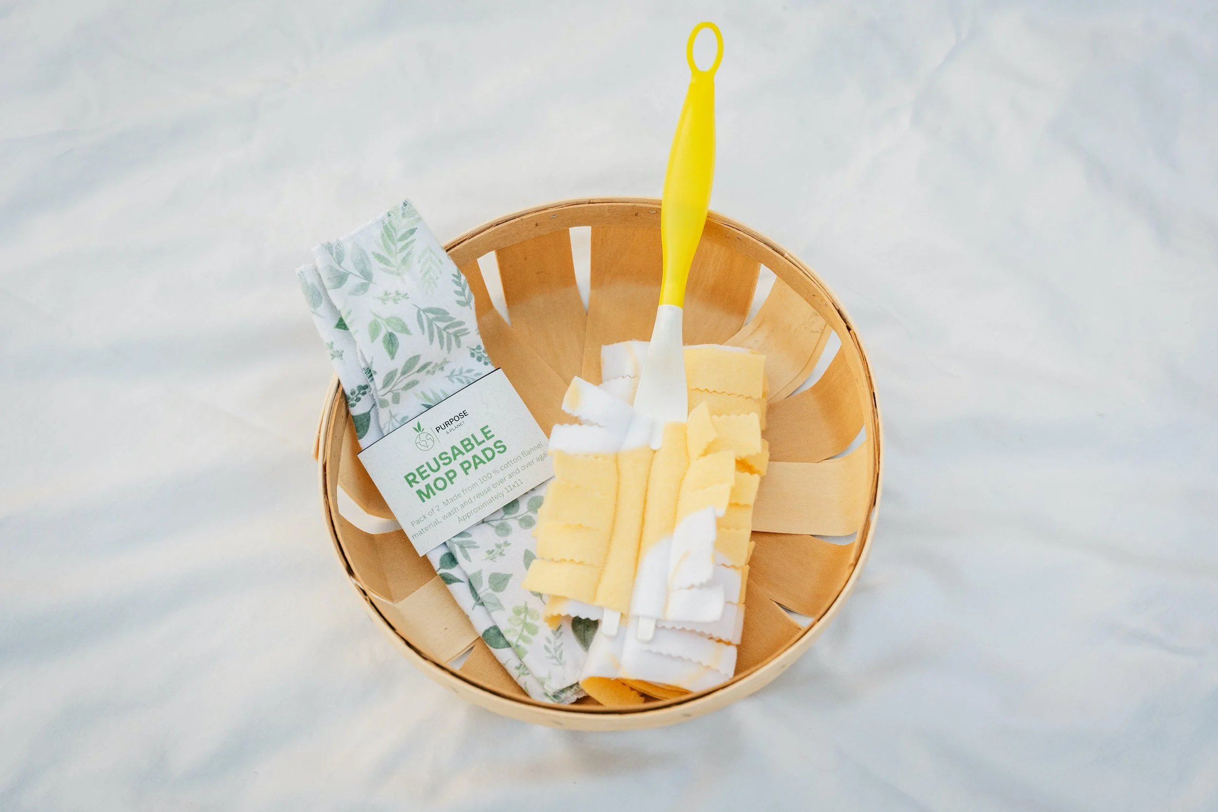 A wooden basket containing reusable mop pads, a dish sponge, and a yellow-handled mop cleaning tool, set on a white surface.