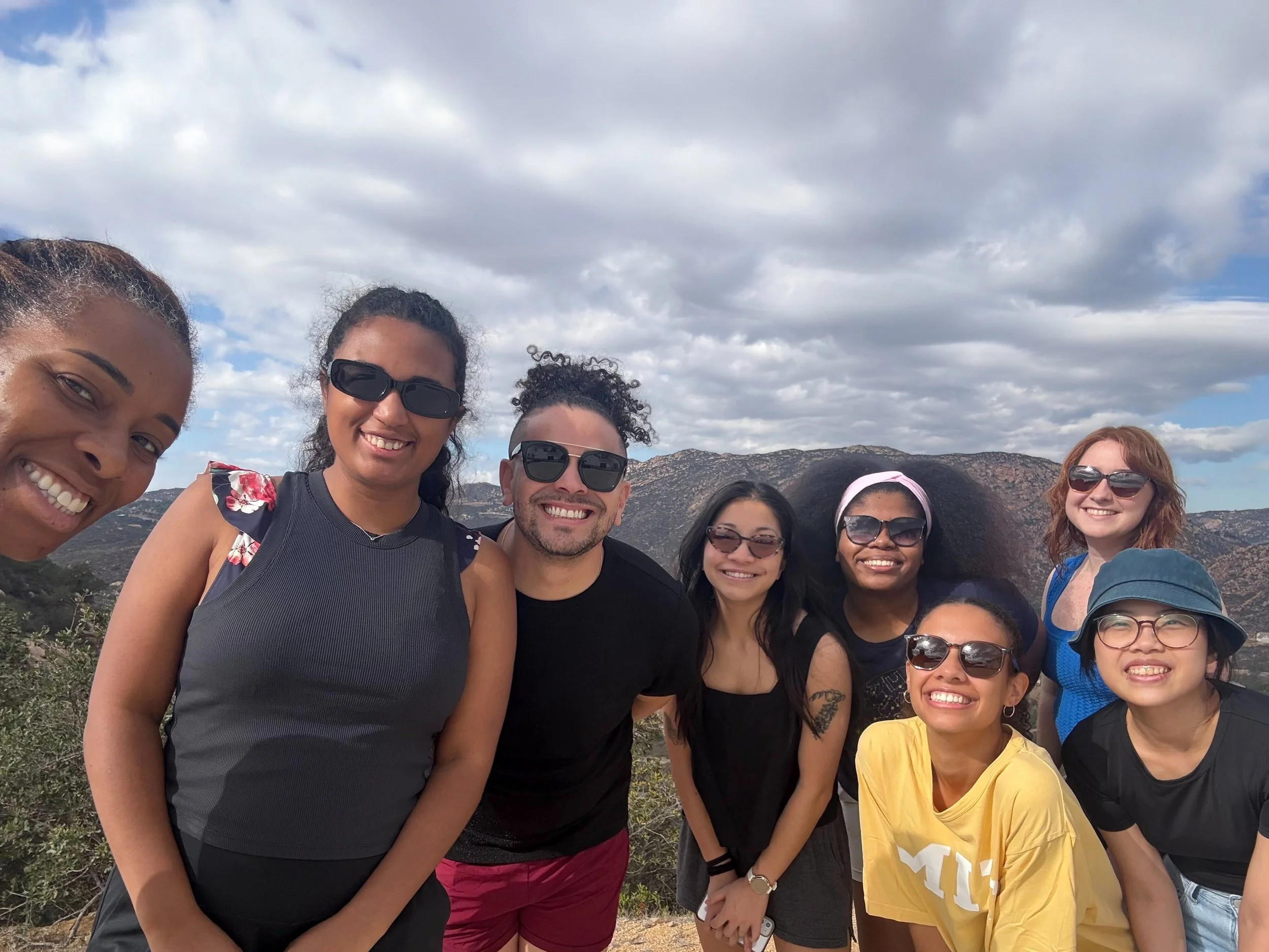 Bianca and her lab members smiling outdoors on a mountain trail under cloudy sky.