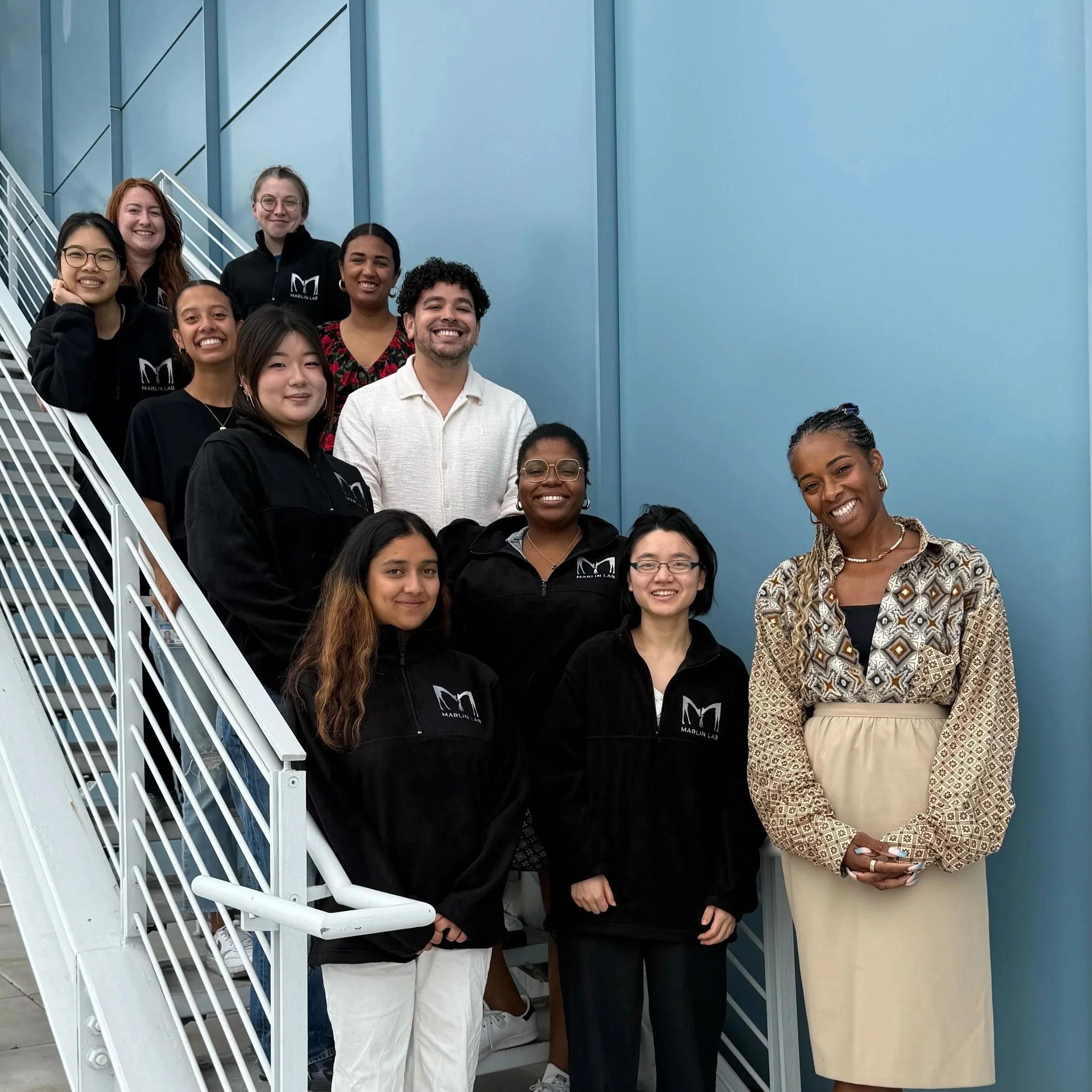 Group of people standing on stairs, smiling, indoors with a plain blue wall background. Some members are wearing black hoodies with the Marlin Lab logo.