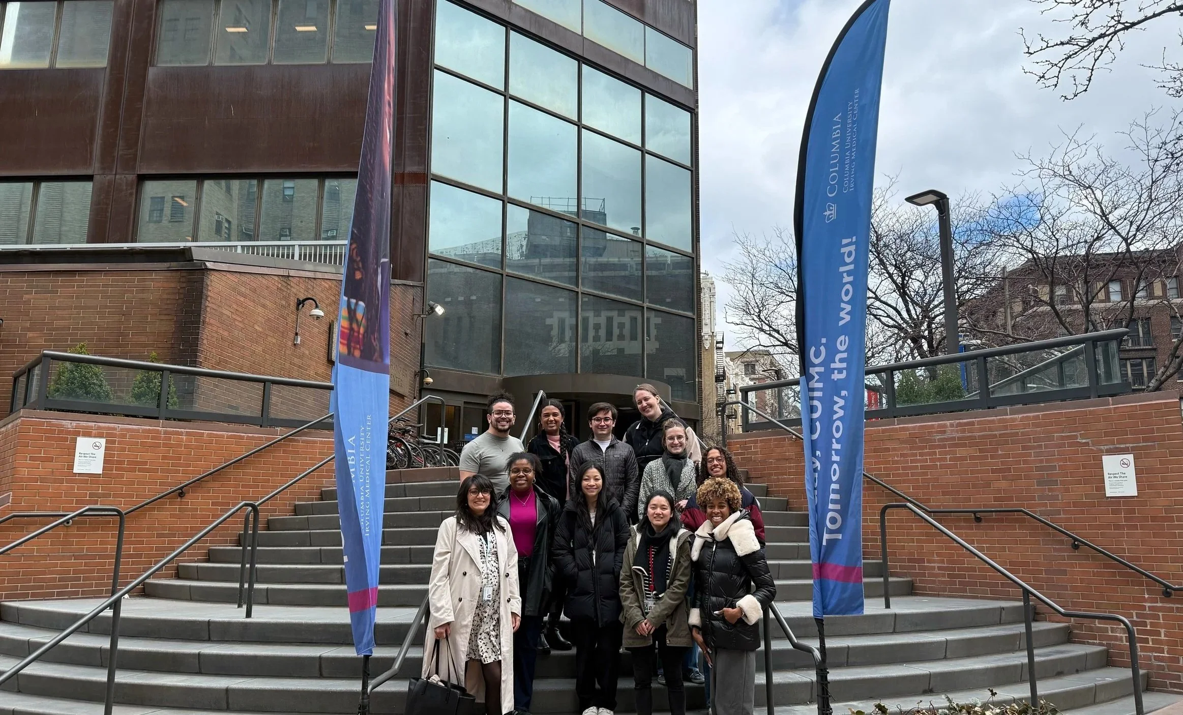 Group of people standing on steps outside Columbia University in New York City during the daytime with flags and modern buildings in the background.