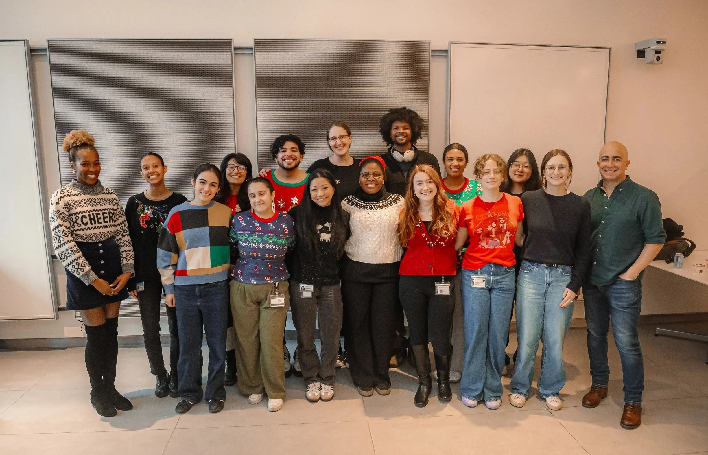 Fifteen people standing in a classroom smiling with some people wearing holiday sweaters.