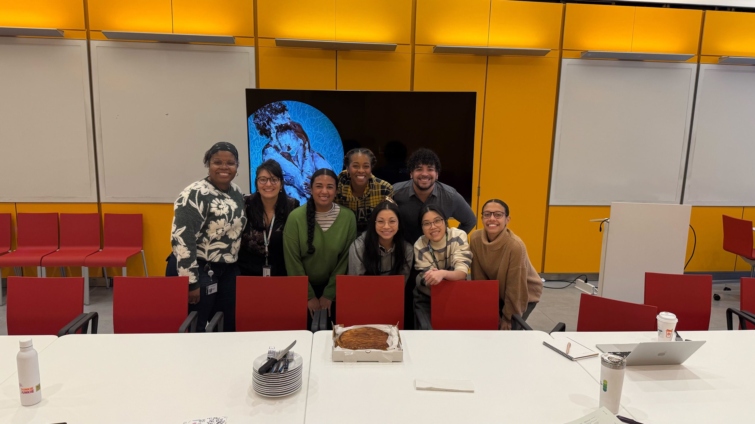 Nine people in a classroom for a celebration in front of a presentation screen with a cake on the table in front of them. 