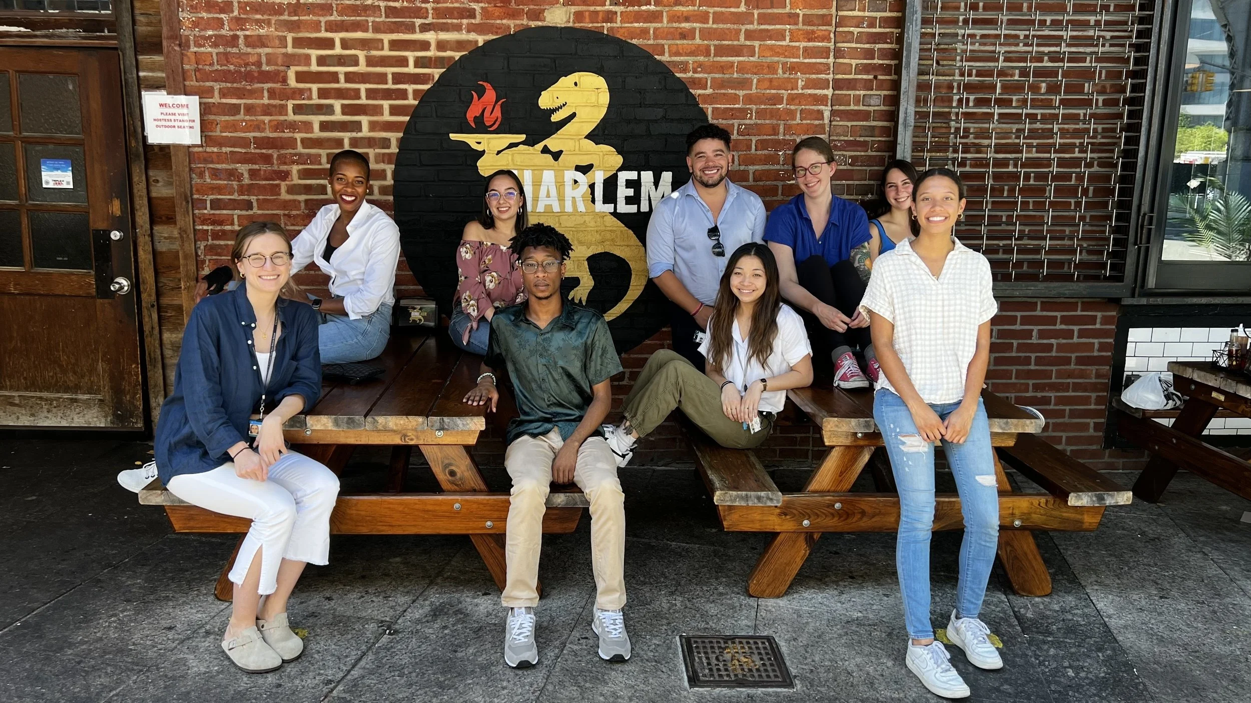 Group of nine young people smiling and sitting on and around a wooden picnic table outside of a brick building with a Harlem Dance Studio sign.
