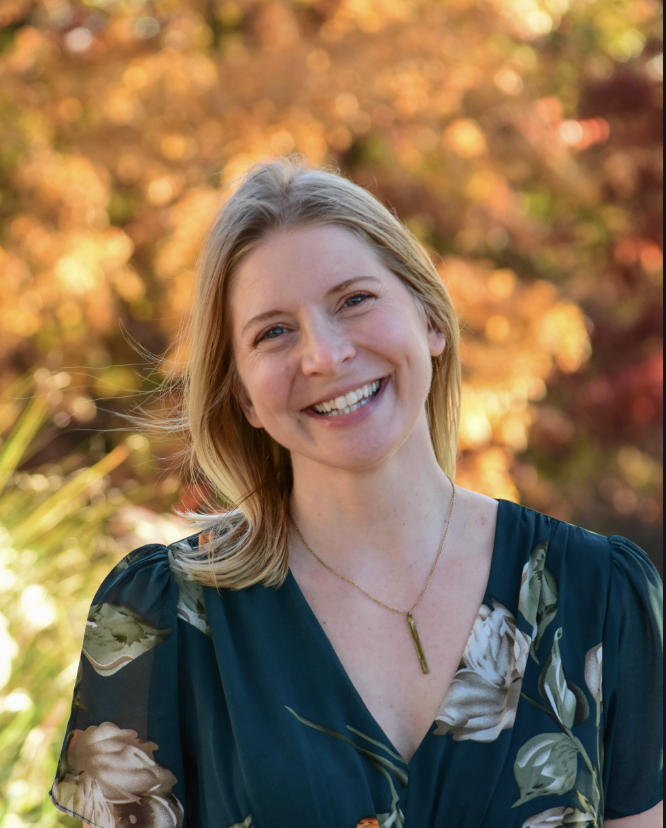 Smiling woman with blonde hair wearing a dark floral blouse outdoors during autumn.