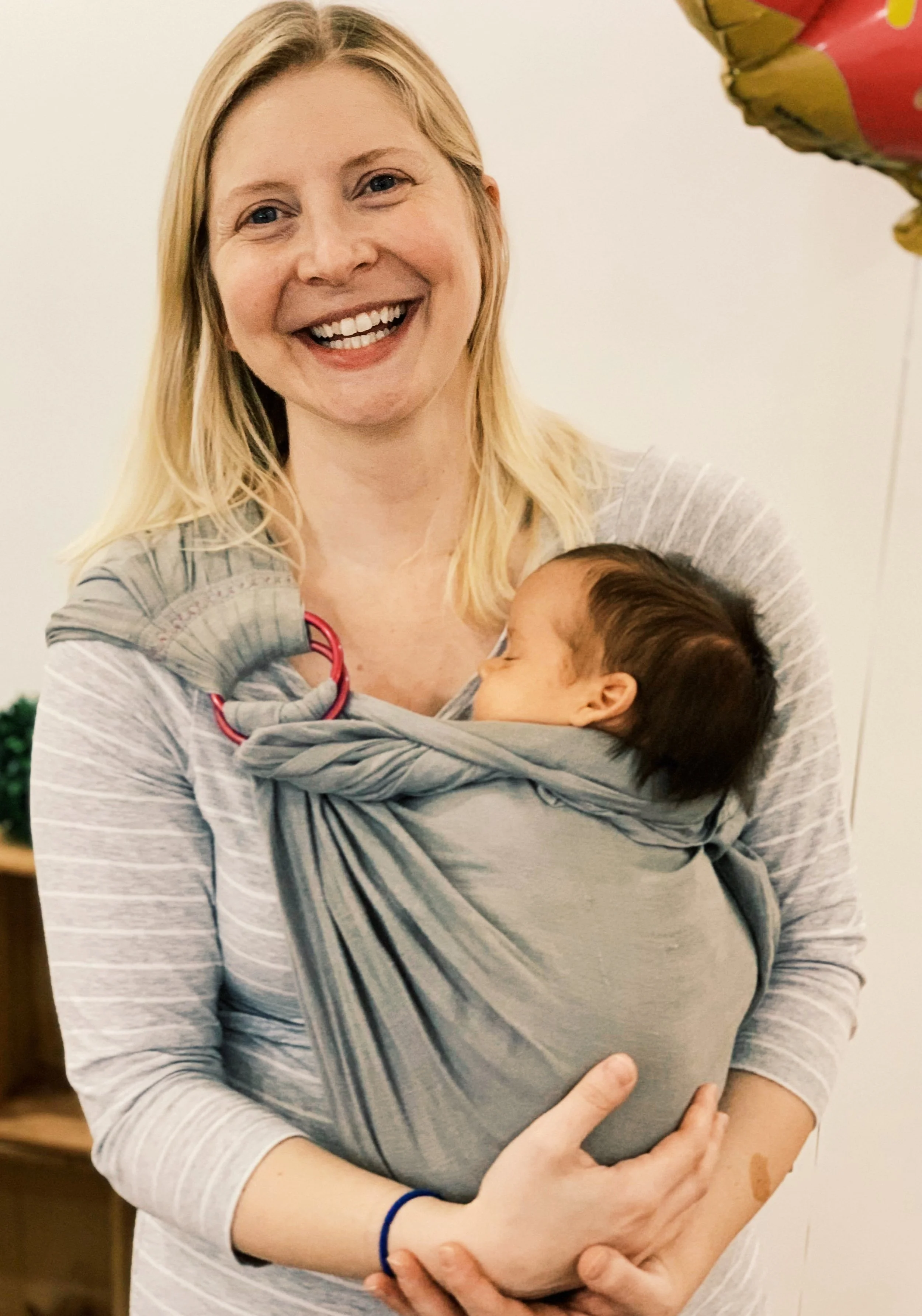 A woman with blonde hair smiling while holding a baby in a baby wrap carrier, with a balloon partly visible in the top right corner.
