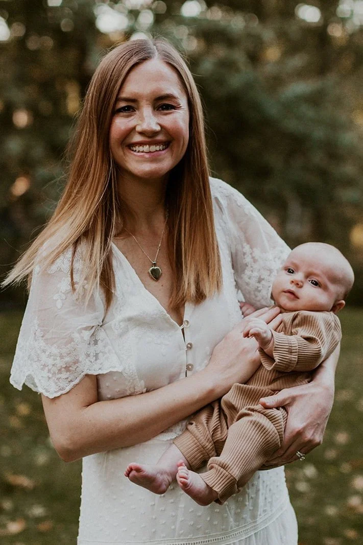 A woman with long, straight, light brown hair smiling and holding a baby outdoors in a park or forest area.