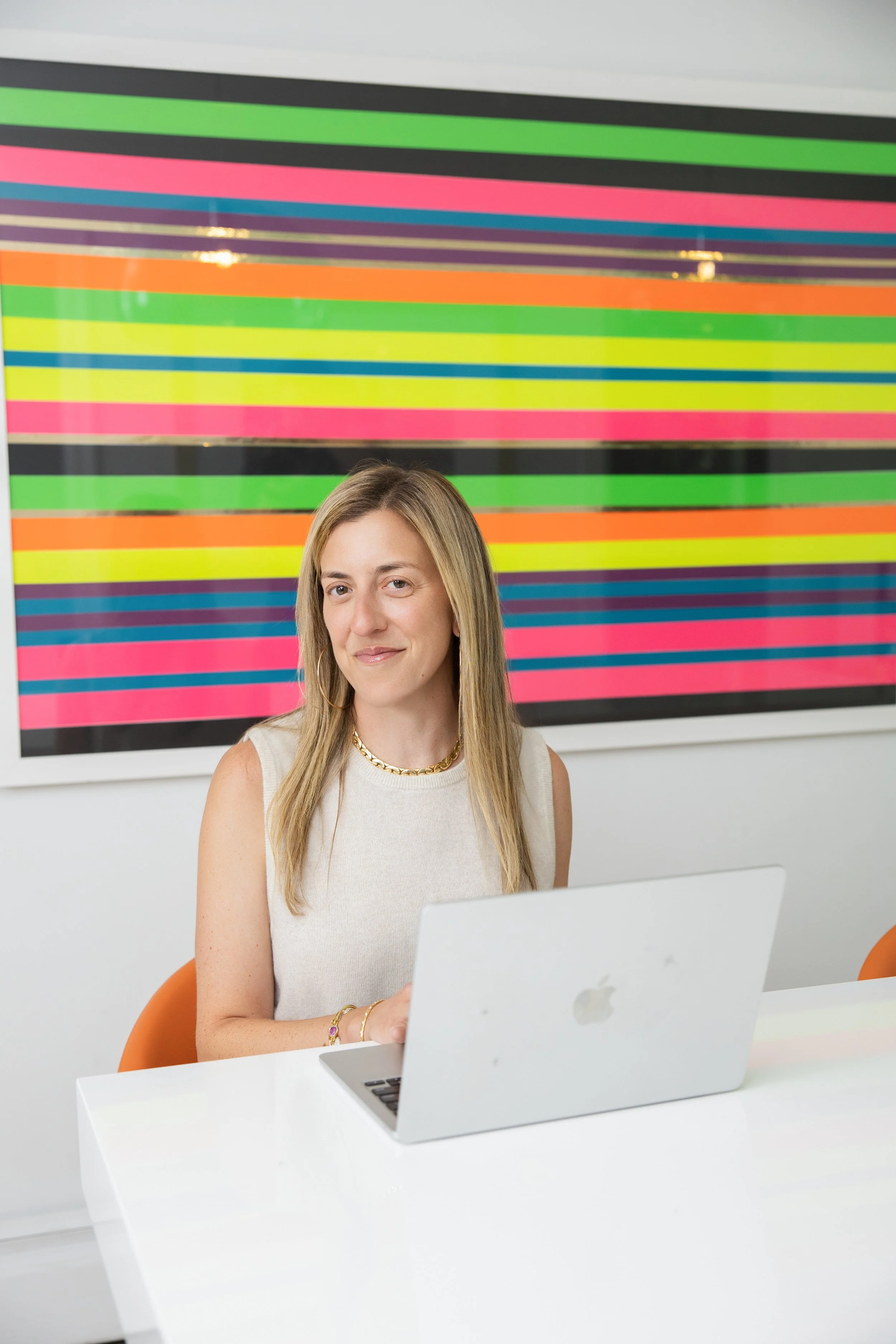 Woman sitting at a white table with a silver MacBook in front of her, smiling mildly, with a colorful striped abstract artwork on the wall behind her.
