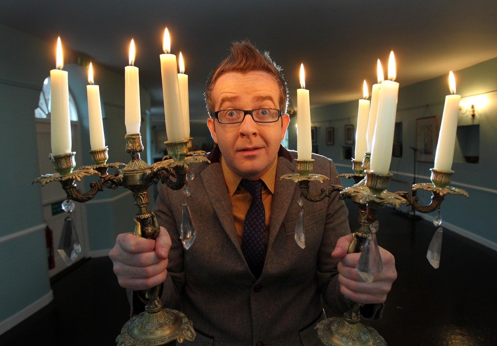 A person wearing glasses and a suit holds two candelabras with lit candles, standing indoors.