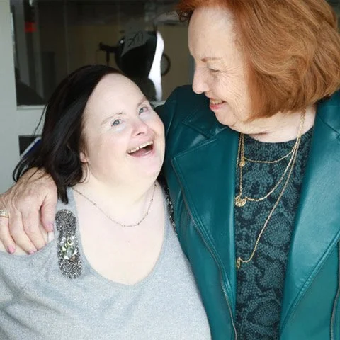 Michelle, an older woman with Down syndrome smiles as she is embraced by her caregiver.
