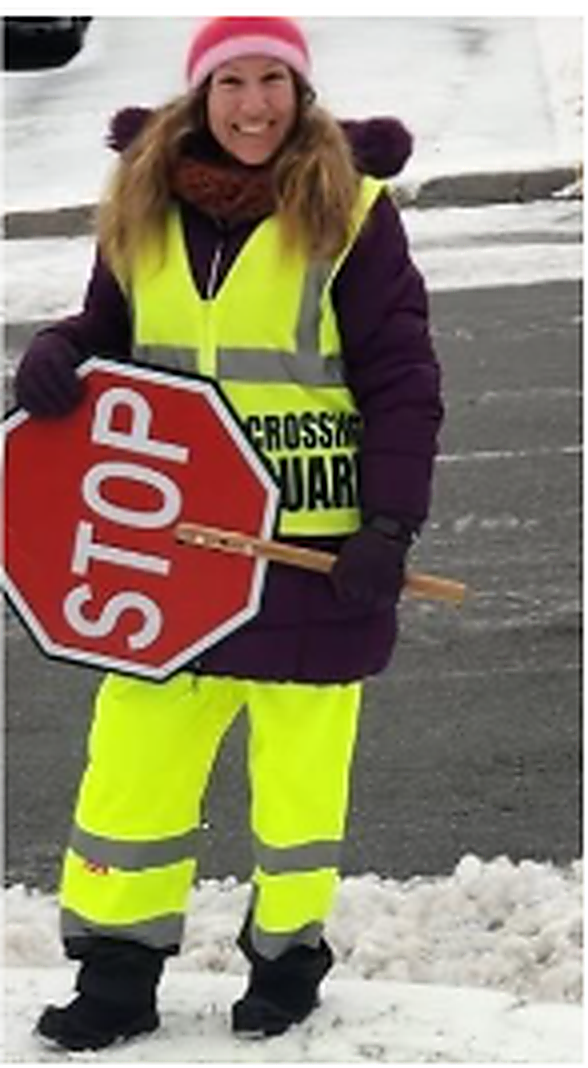 A woman wearing a fluorescent yellow safety vest and pants stands outdoors in winter holding a red STOP sign and a crossing-guard paddle.