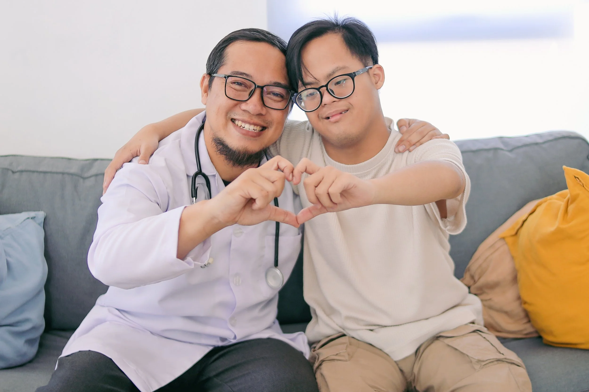 A doctor with dark hair, glasses and a white coat with a stethoscope around his neck, has an arm around a young adult with down syndrome wearing glasses. Both are making a heart shape with their hands.