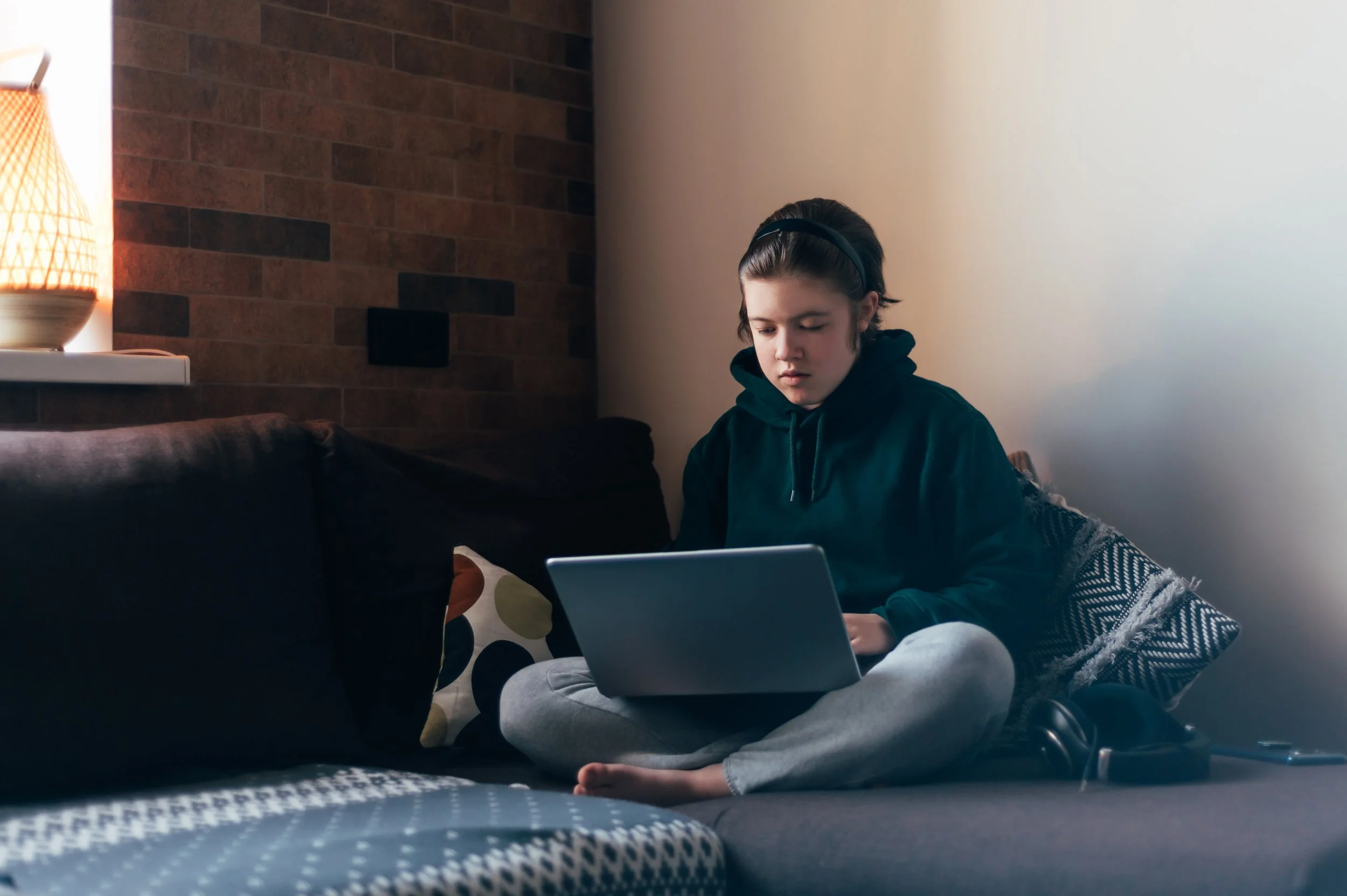 A woman with short hair and a dark green hoodie is seated cross-legged on a gray sofa while looking at a laptop. The scene is indoors, in a room with brick-like wall paneling. A lamp sits on a windowsill behind the sofa.