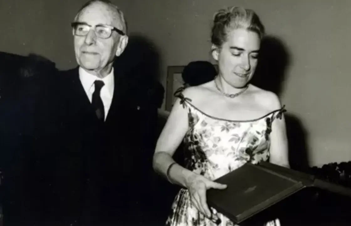 She wears a shoulder-baring flower-print dress while holding an award folder. Her hair is swept back off her head.