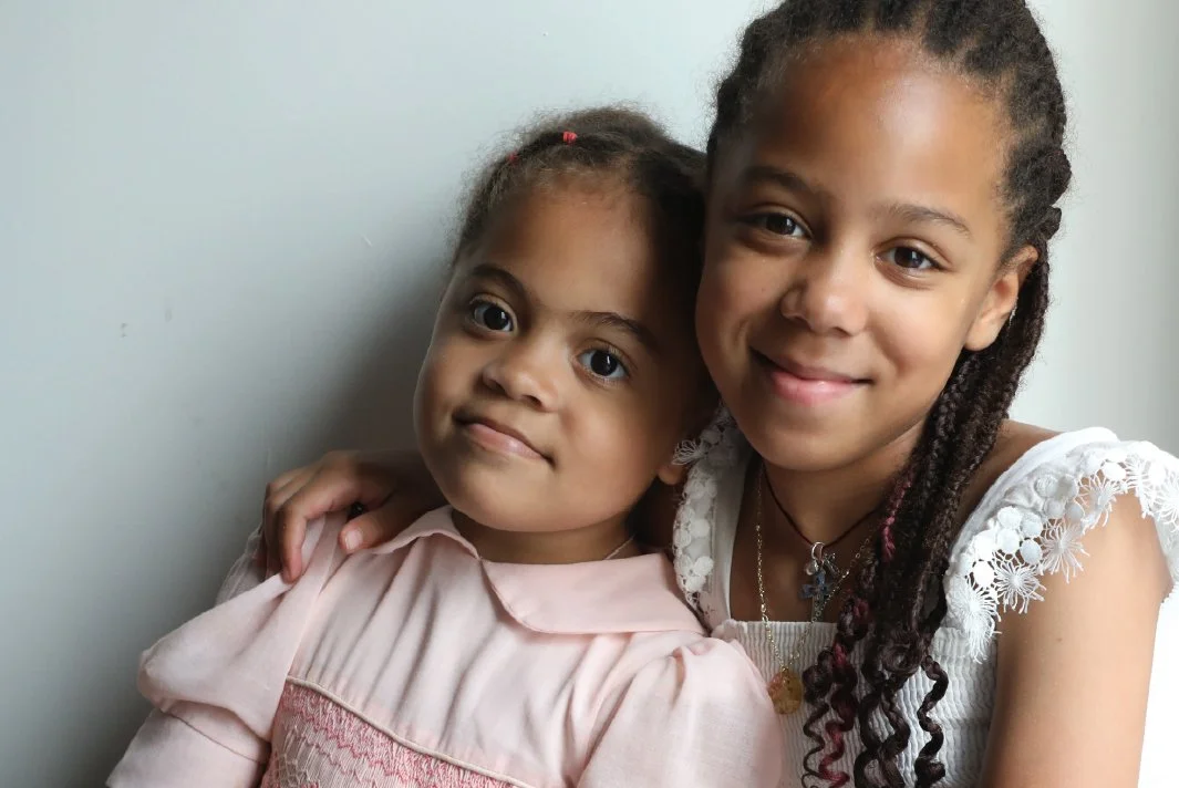 Sophia, in a sweet pink dress leans against Olivia, dressed in pretty white lace