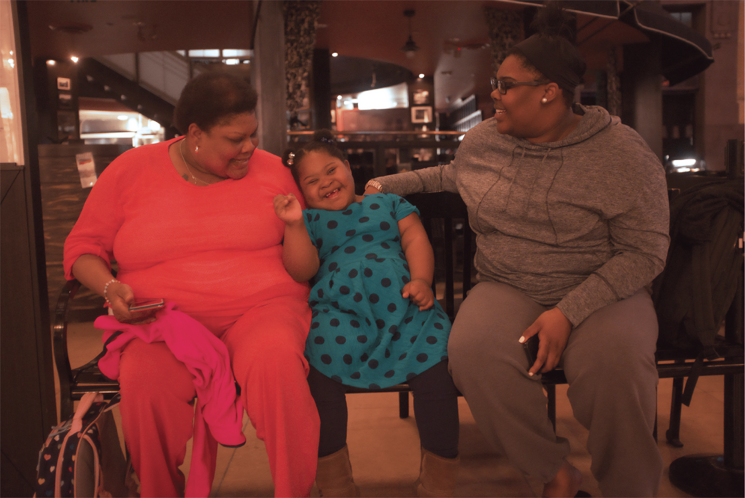 A child with Down syndrome giggles as she sits between her mother and grandmother on a bench.