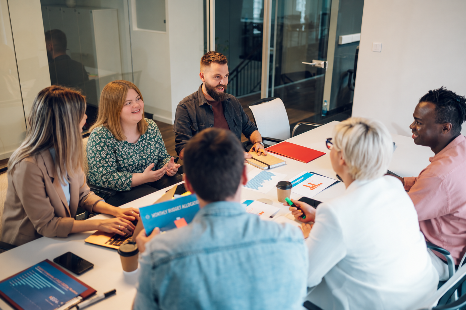 A team of six adults, including men and women, are collaborating in a bright, modern office setting. They are gathered around a table, reviewing charts, graphs, and other documents. The atmosphere appears professional and focused.
