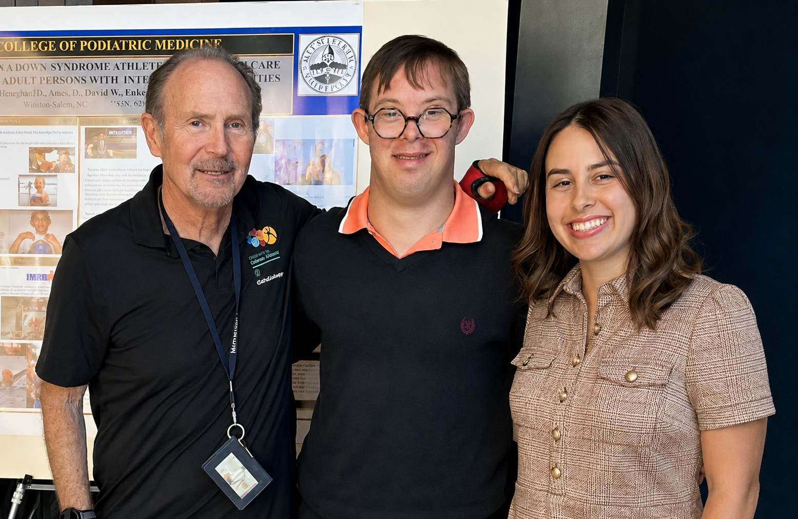 Three people stand together in front of an academic poster about Down syndrome, athletics, and healthcare.